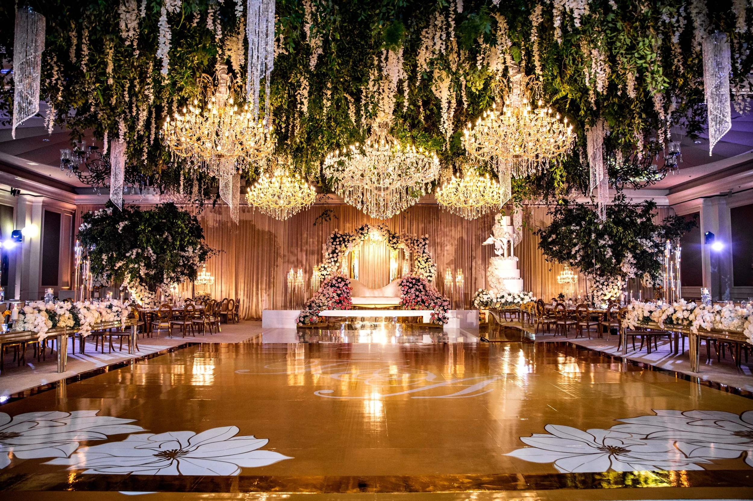 A lavish wedding reception hall decorated with chandeliers, flowers, and elegant furnishings, featuring a dance floor with floral patterns, a floral backdrop behind a wedding chaise lounge, and botanical arrangements.”