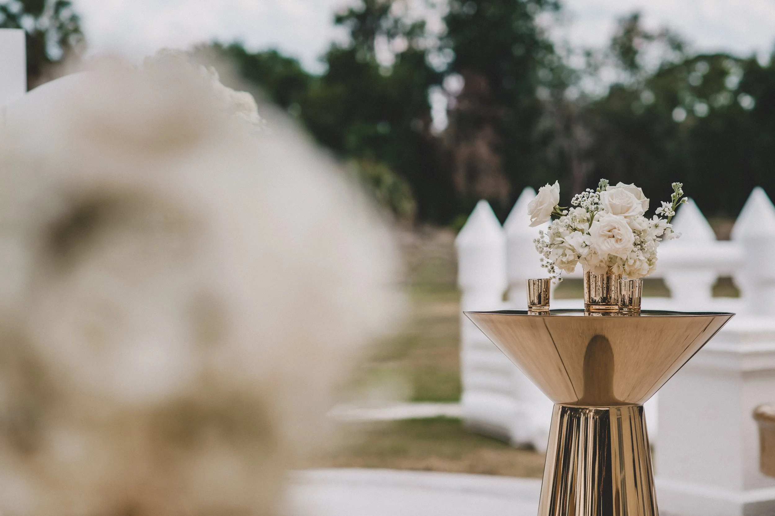White floral arrangement with roses and baby's breath in a gold vase on a gold table at an outdoor event with white tents in the background.