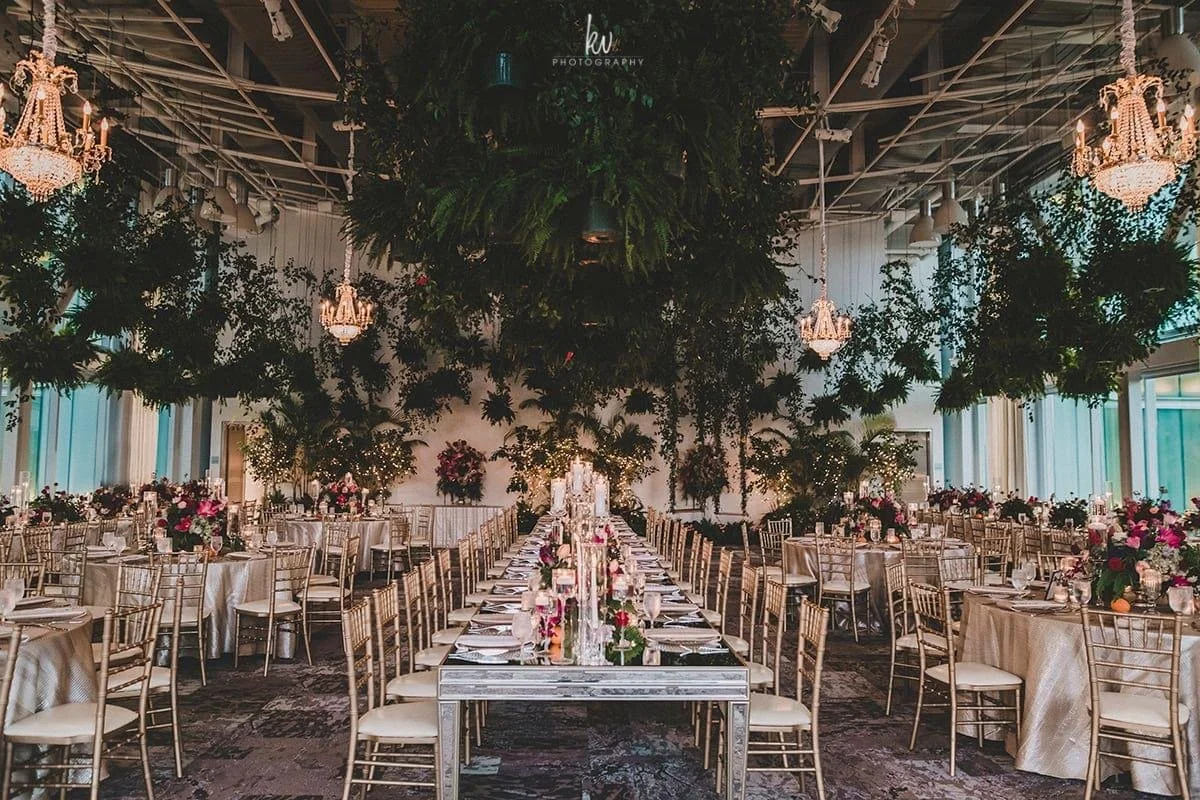 Elegant banquet hall decorated with hanging green foliage and chandeliers, featuring long dinner tables with floral centerpieces and gold chairs.