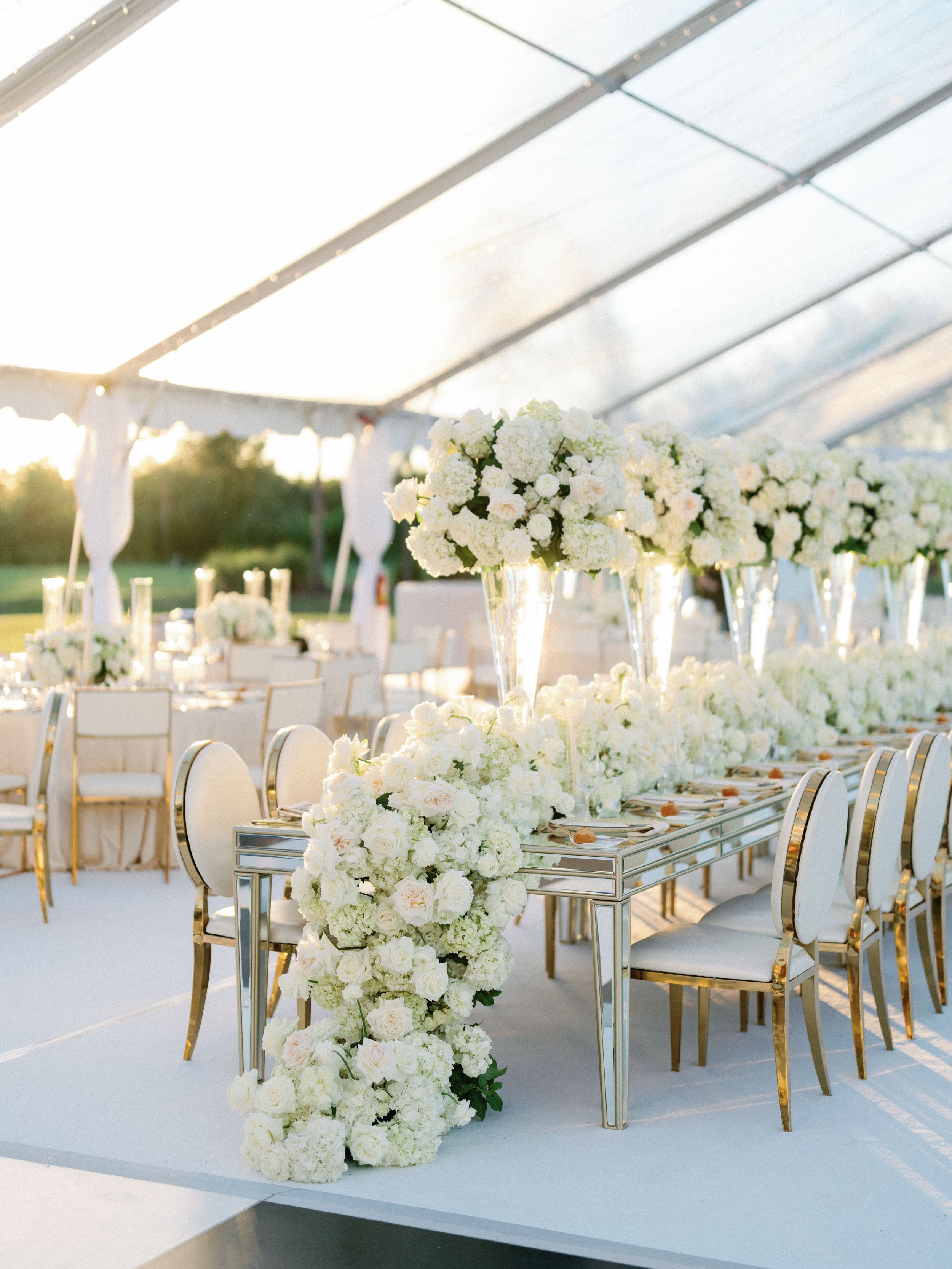 Elegant outdoor wedding reception with long table decorated with white flowers and high floral centerpieces under a clear tent, sunlight glowing in the background.
