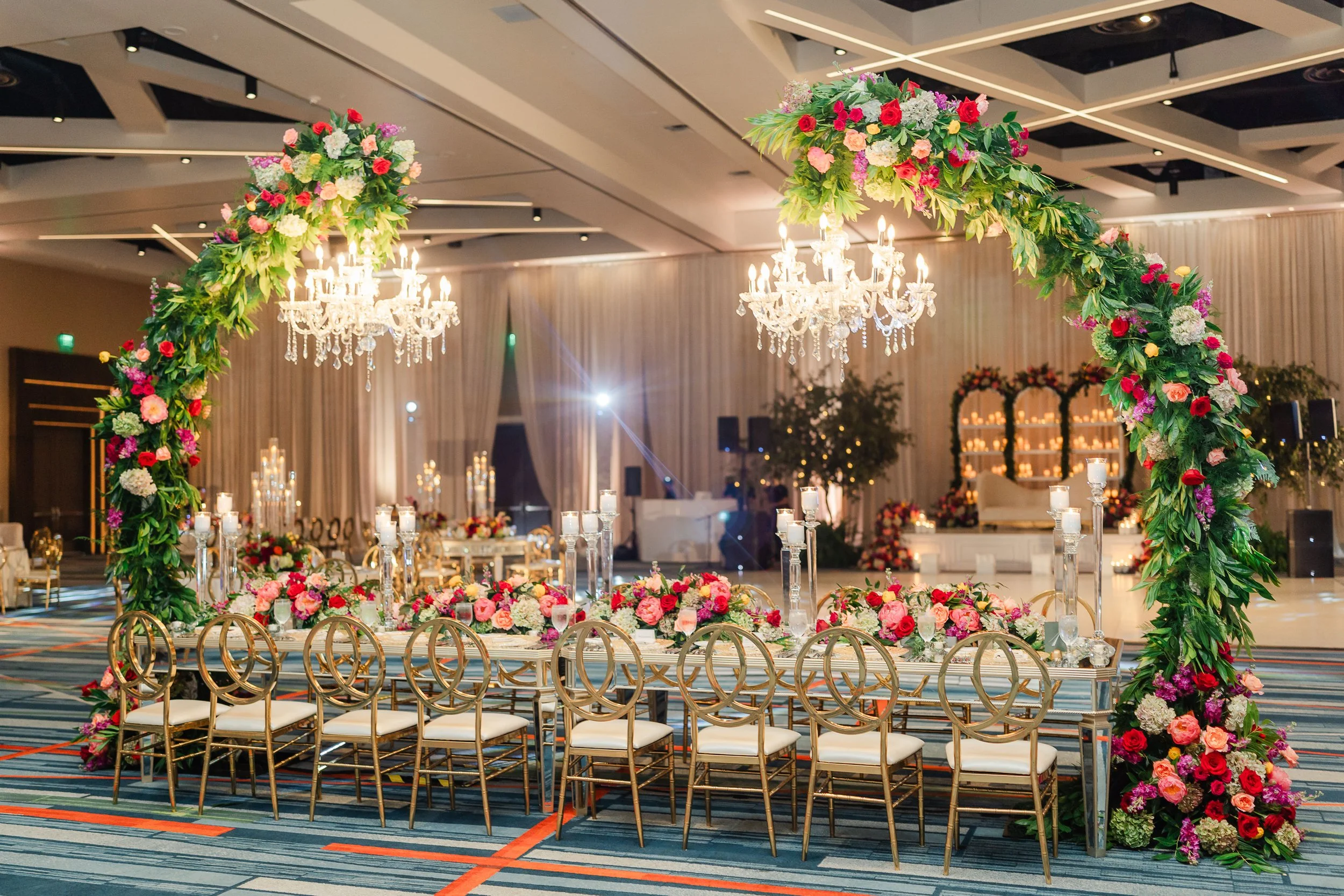 Elegant wedding reception setup with a floral arch, a long table decorated with pink, red, and white flowers, and golden chairs in a decorated ballroom with chandeliers and candlelit displays.