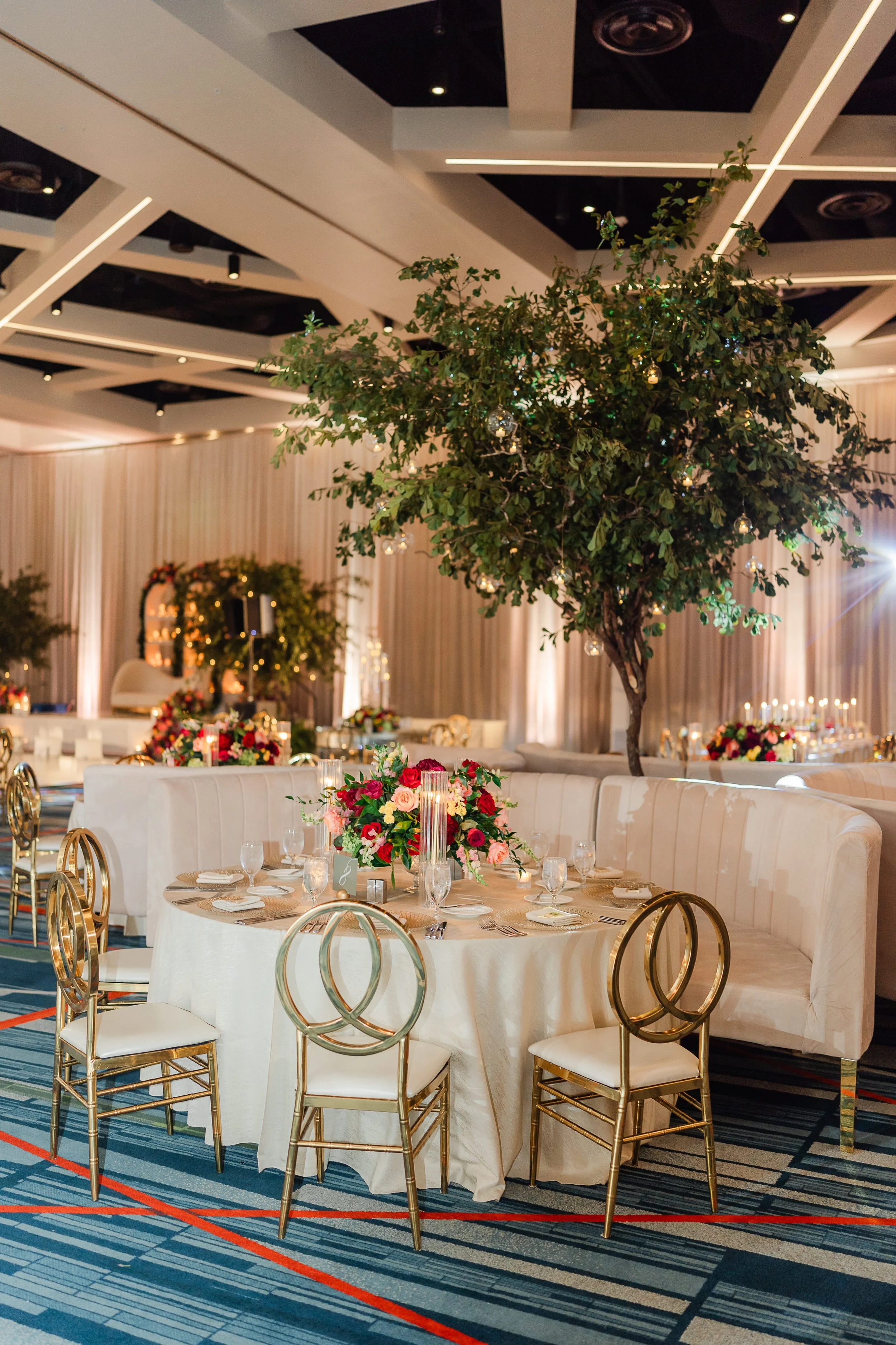 Elegant banquet hall decorated with large floral arrangements, round tables with cream-colored tablecloths, gold chairs, and a large indoor tree with hanging ornaments.