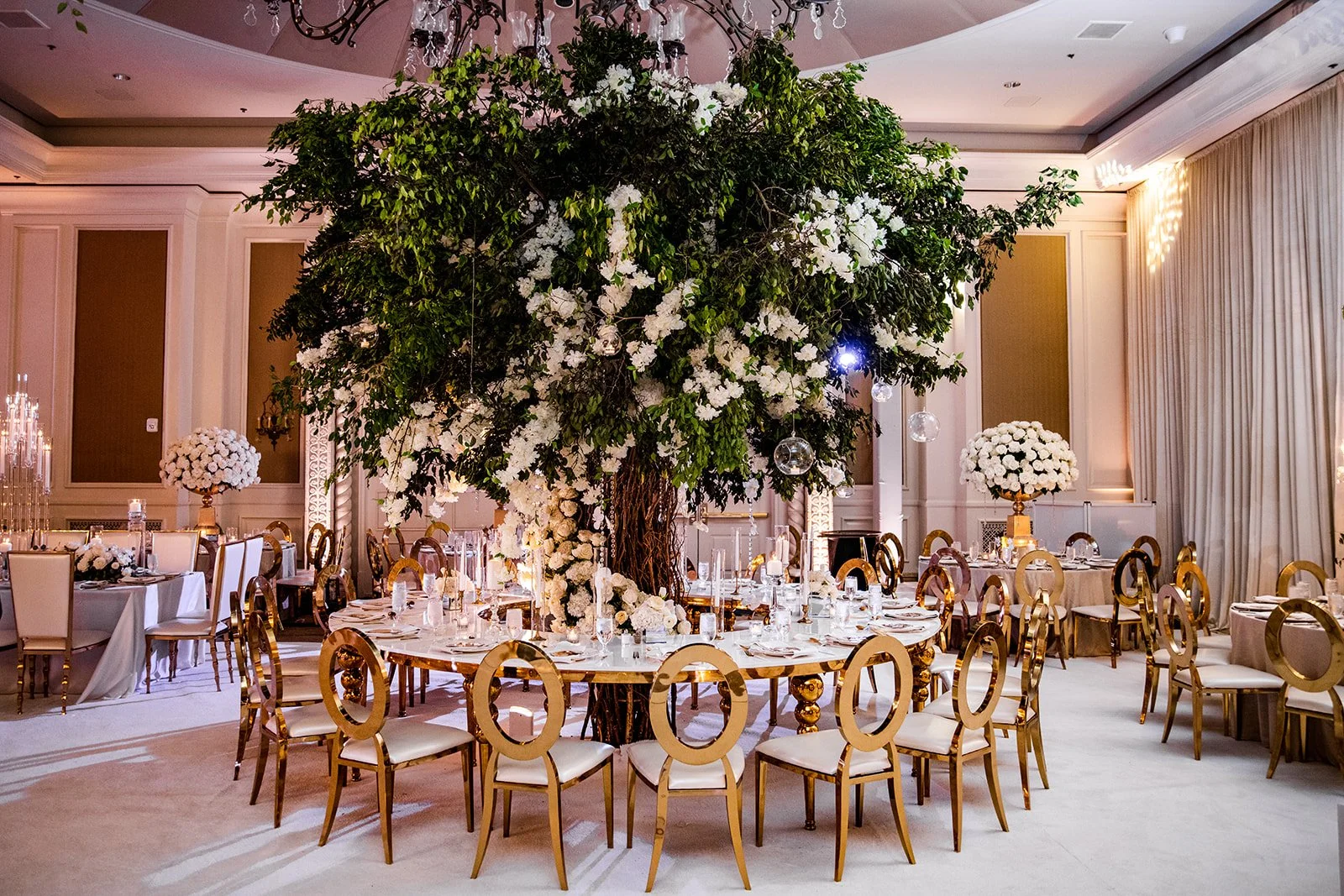 Elegant banquet hall decorated for an event with a large tree centerpiece adorned with white flowers and hanging glass ornaments, surrounded by round tables with white tablecloths, floral centerpieces, and gold chairs.