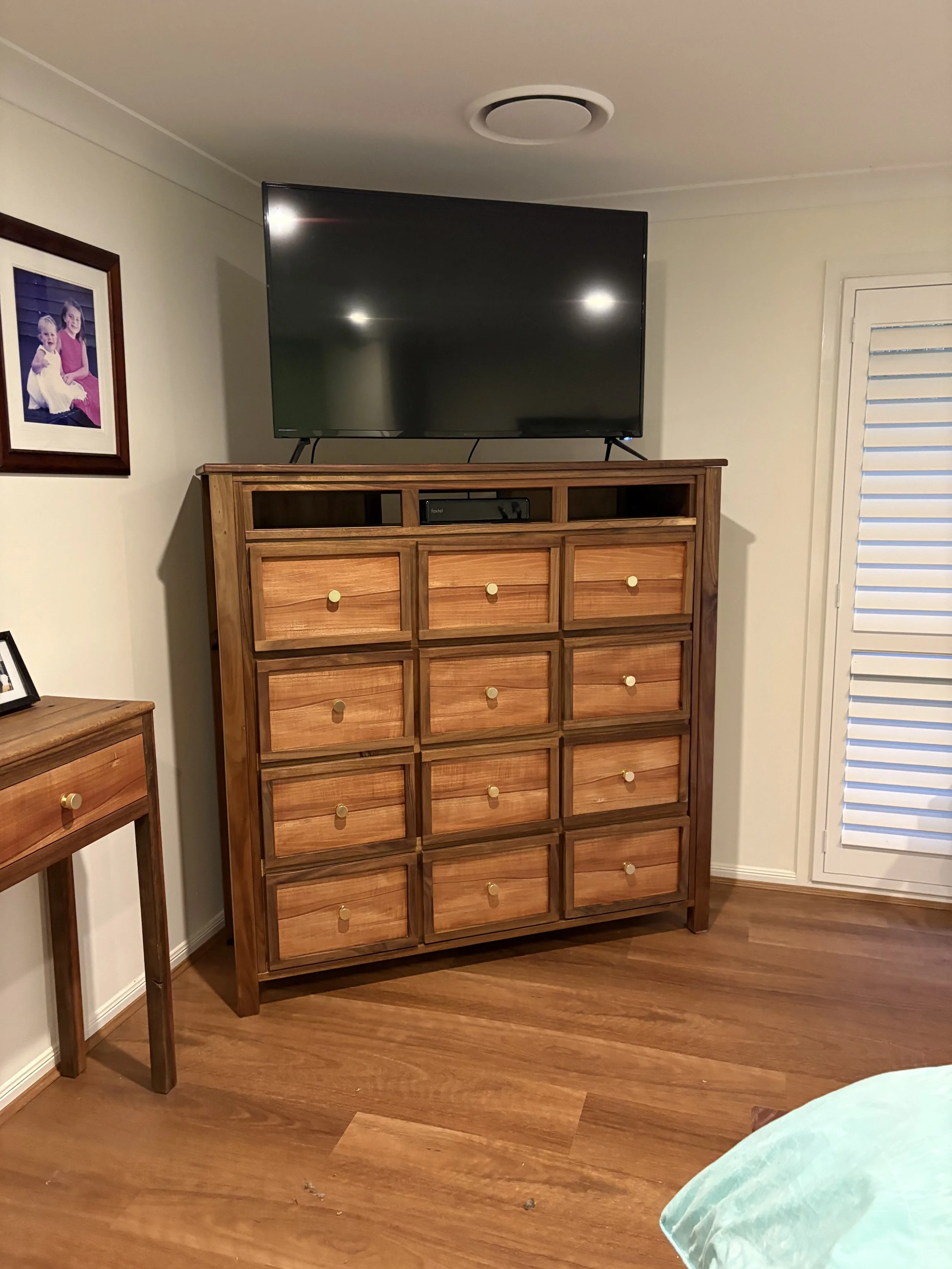 A flat-screen TV on top of a wooden dresser with multiple drawers in a bedroom. There is a framed family photograph on the wall to the left and a small wooden table partially visible on the left. A door with horizontal louvered panels is on the right.