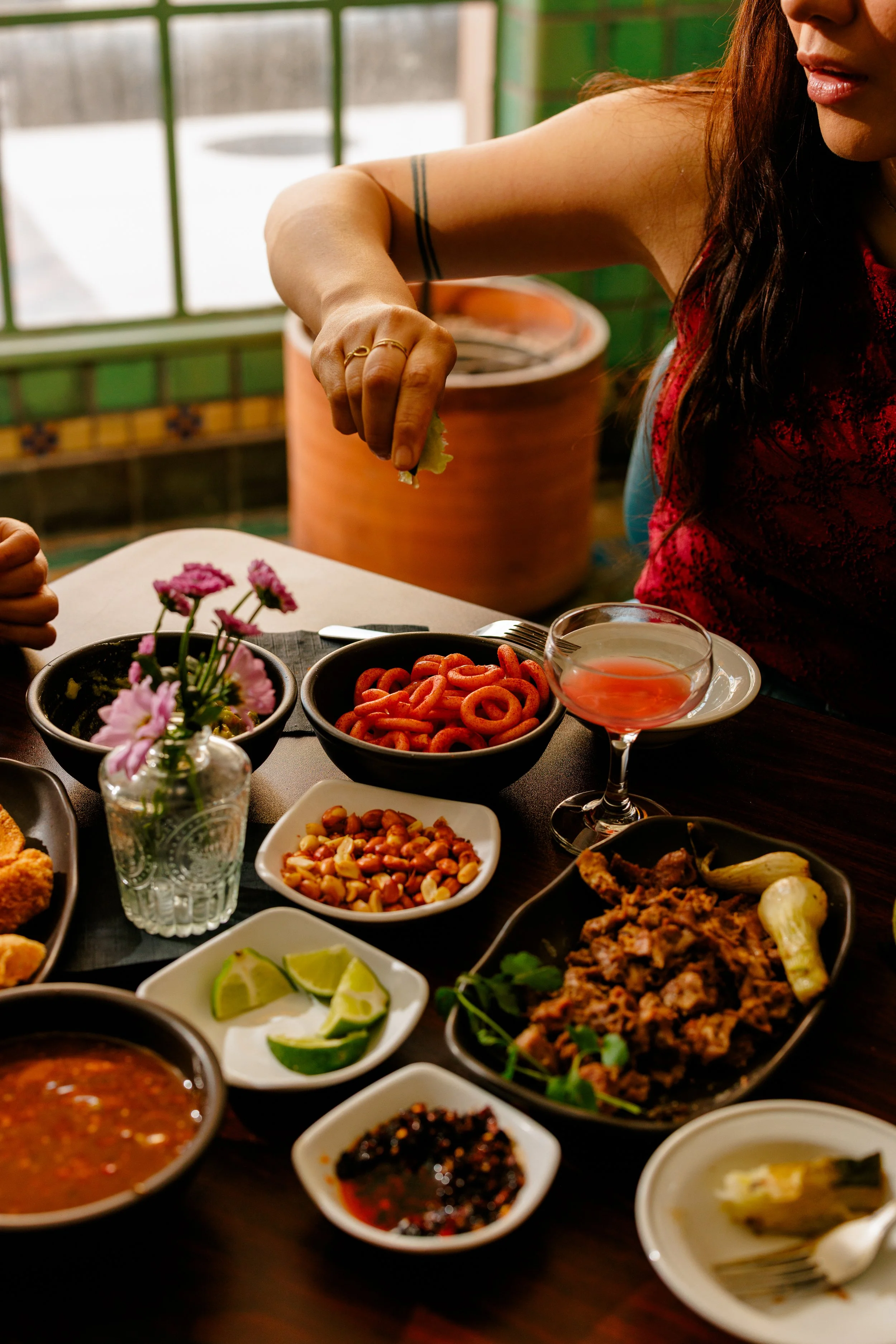Person squeezing lime over a variety of Mexican dishes on a table, including salsa, jalapeños, and tortilla chips, with a potted plant and window in the background.