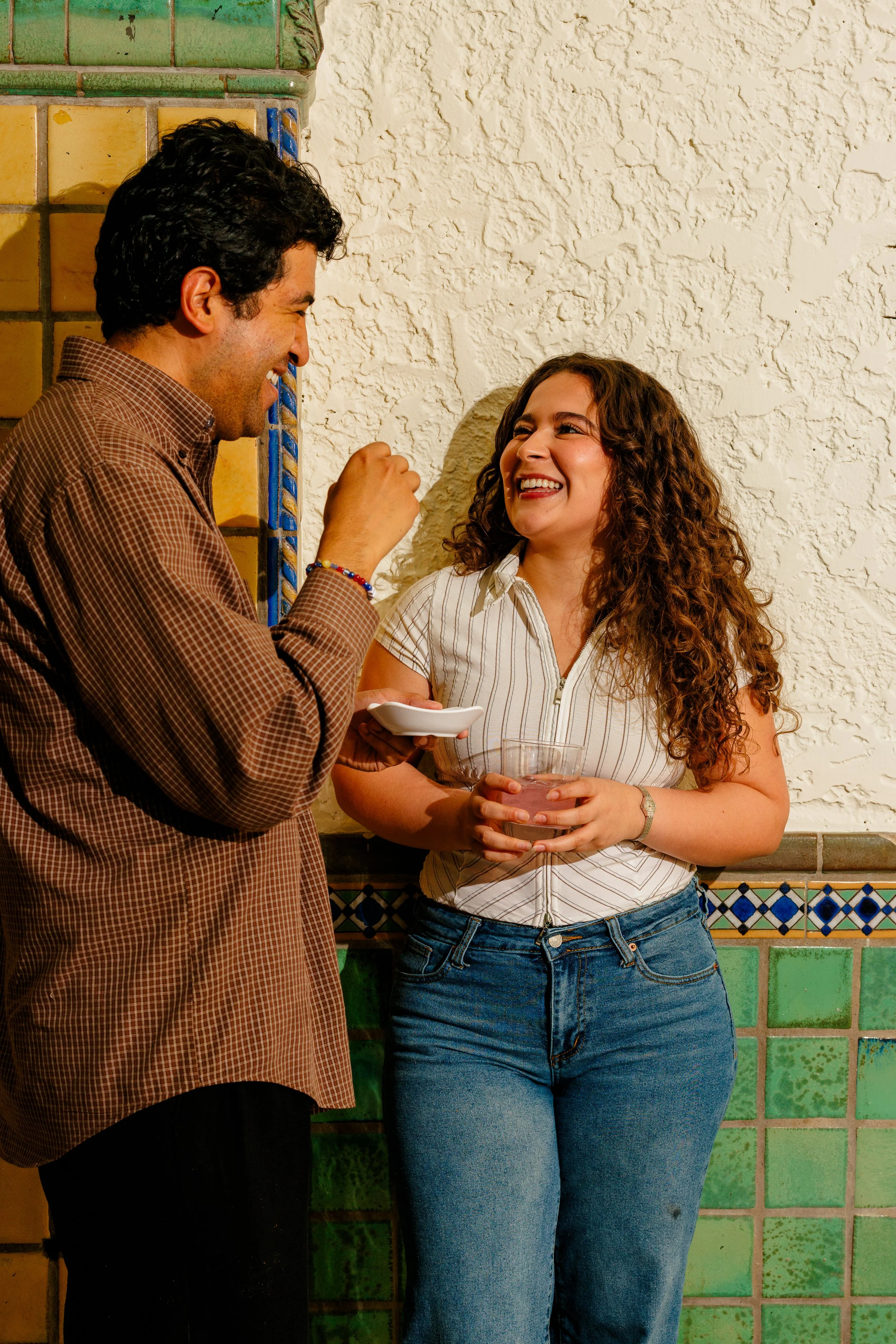 A man and woman laughing and talking, standing against a tiled wall.