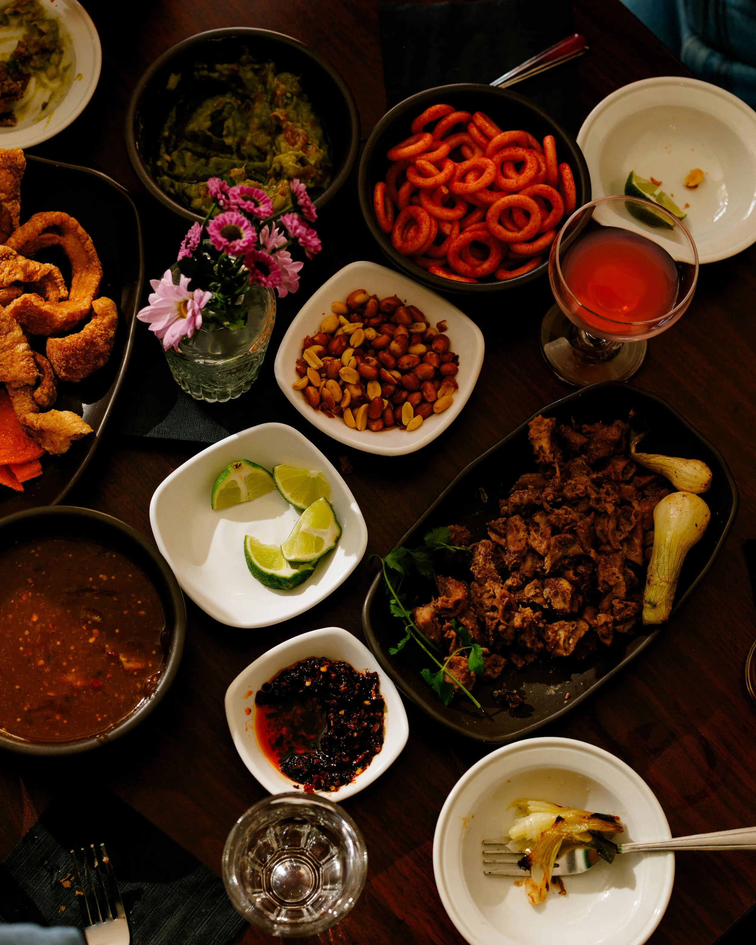 Assorted dishes and snacks on a wooden table, including lime wedges, chili paste, fried onion rings, peanuts, spiral pasta, grilled meat with garlic, and a glass of pink beverage.