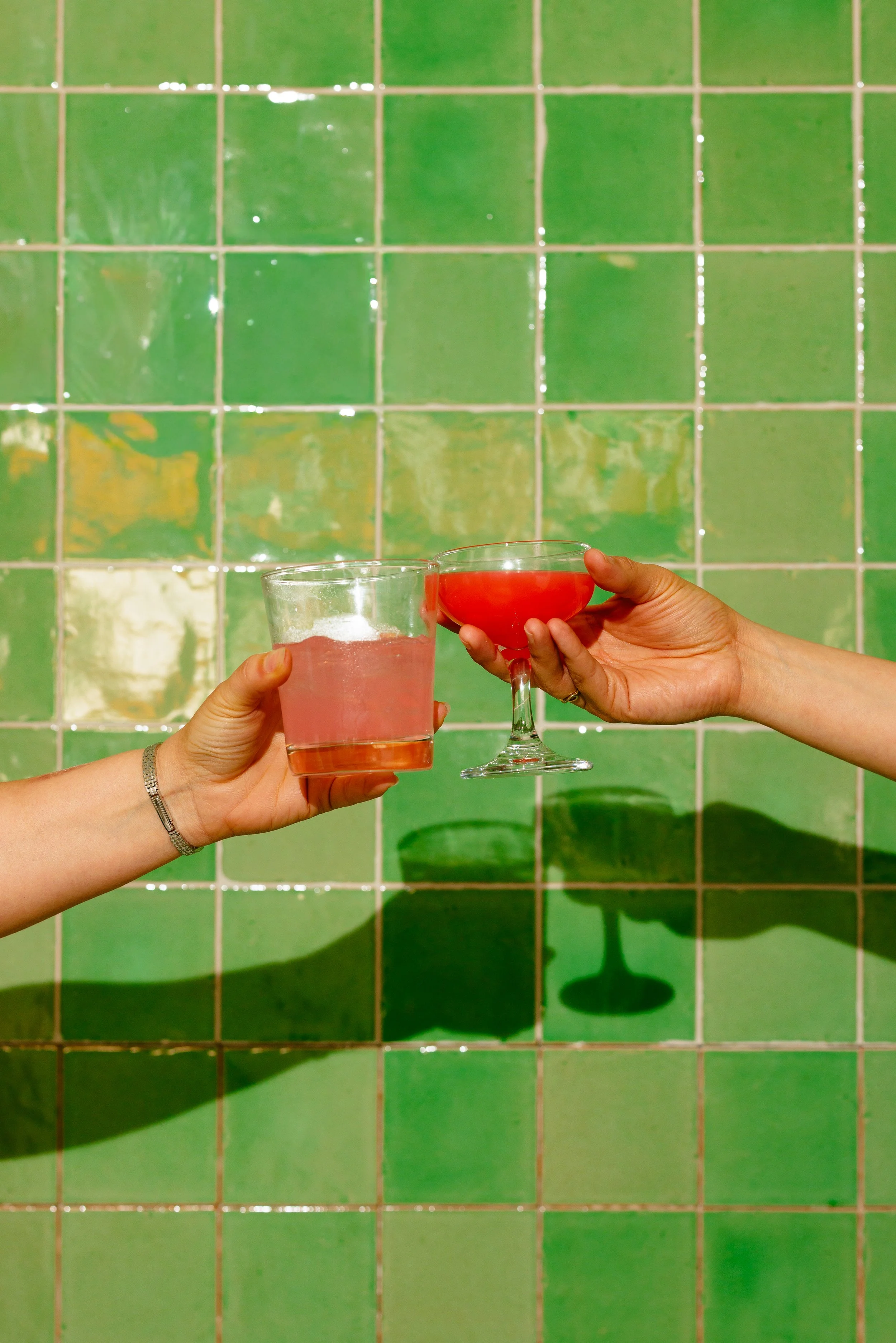 Two hands toast with glasses of pink and red beverages against a green tiled wall.