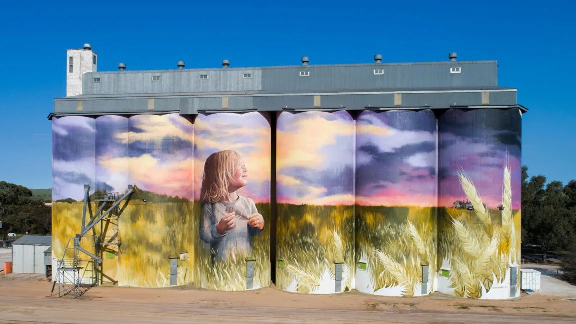 A silo with a mural of a young girl in a field with a sunset sky painted on it.
