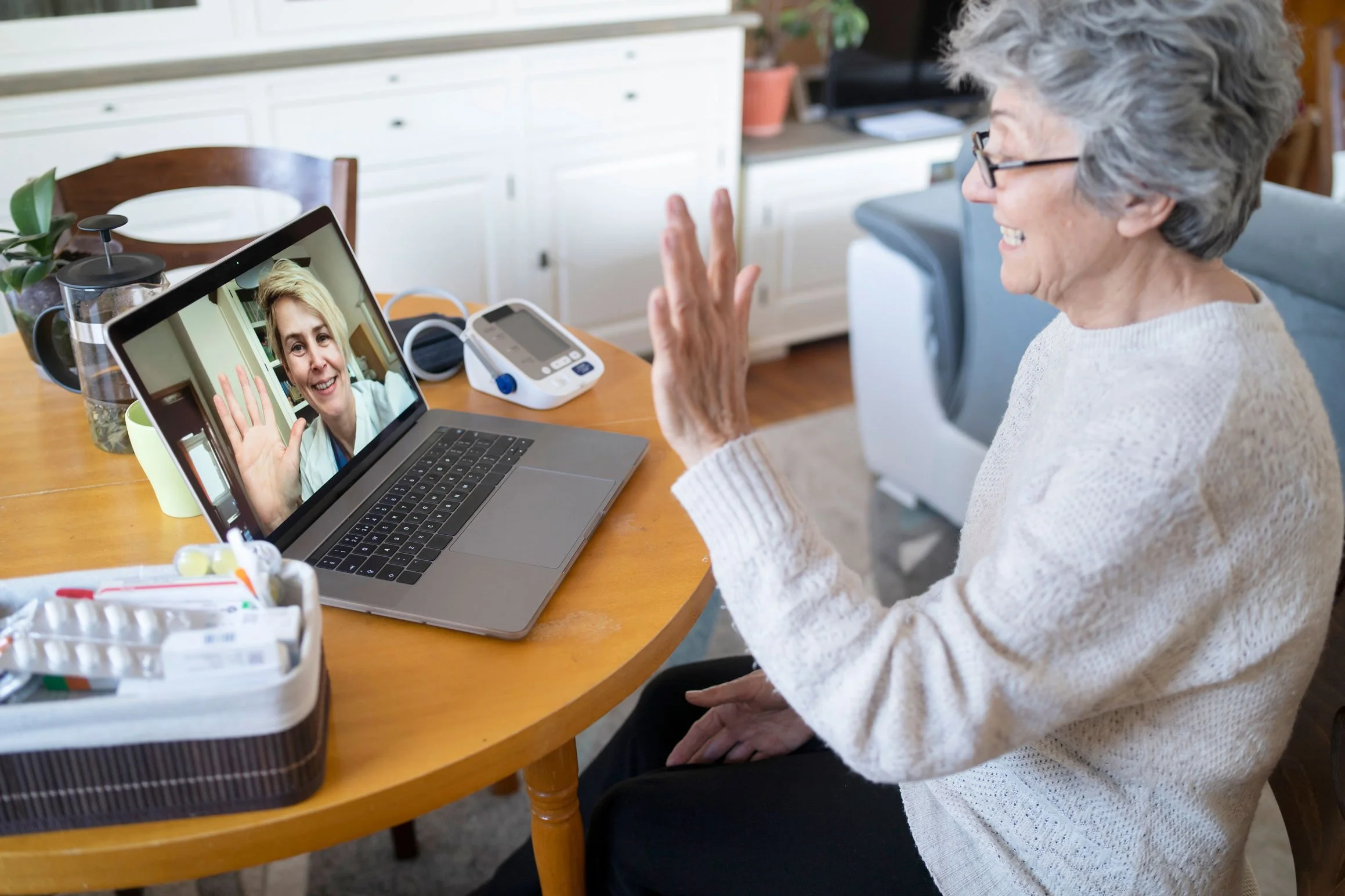 An elderly woman with gray hair and glasses is talking to a woman on a video call, waving and smiling on her laptop screen. The elderly woman is seated at a wooden table with medical items and plants around, in a bright kitchen or dining area.