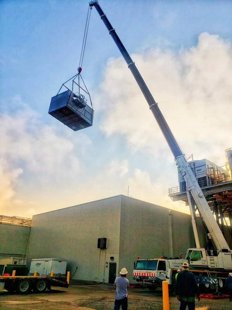 Crane lifting a large industrial container above a warehouse with several workers observing.