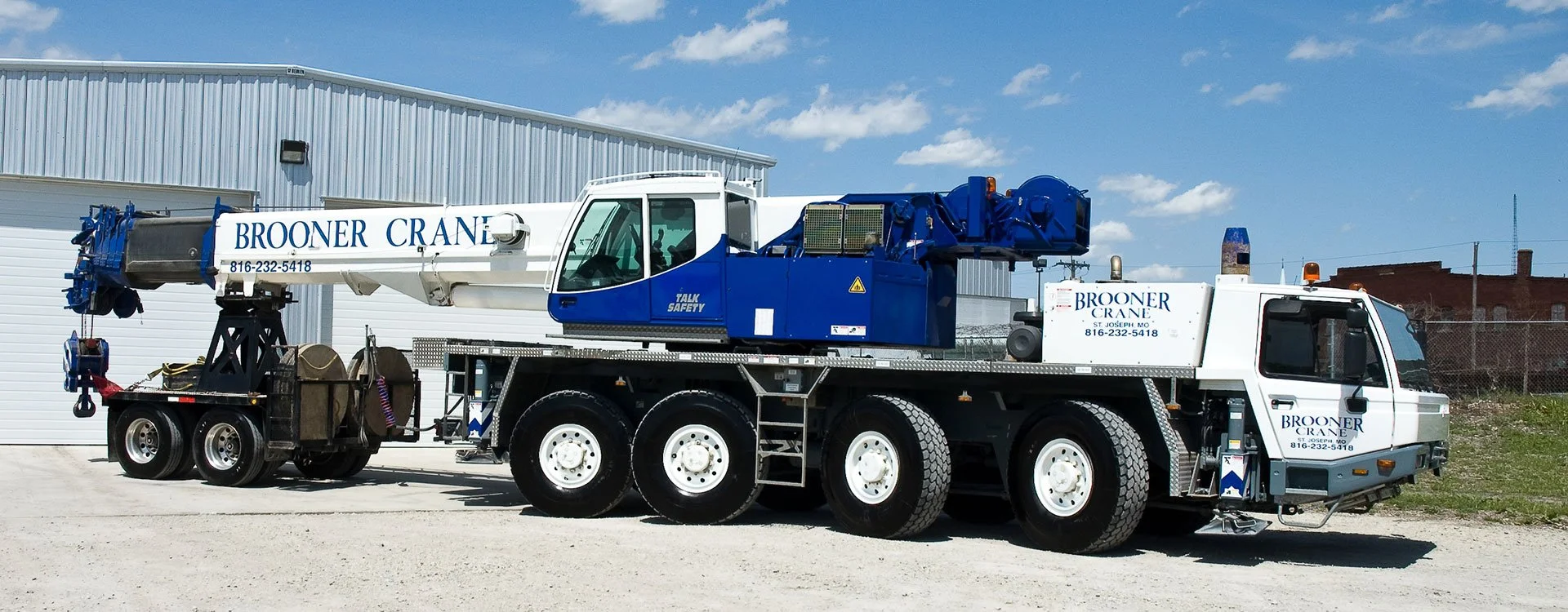 A large white and blue crane truck with the company name 'Brooner Crane' and phone number on the arm and side, parked on a gravel lot near a warehouse with a chain-link fence and a clear blue sky.