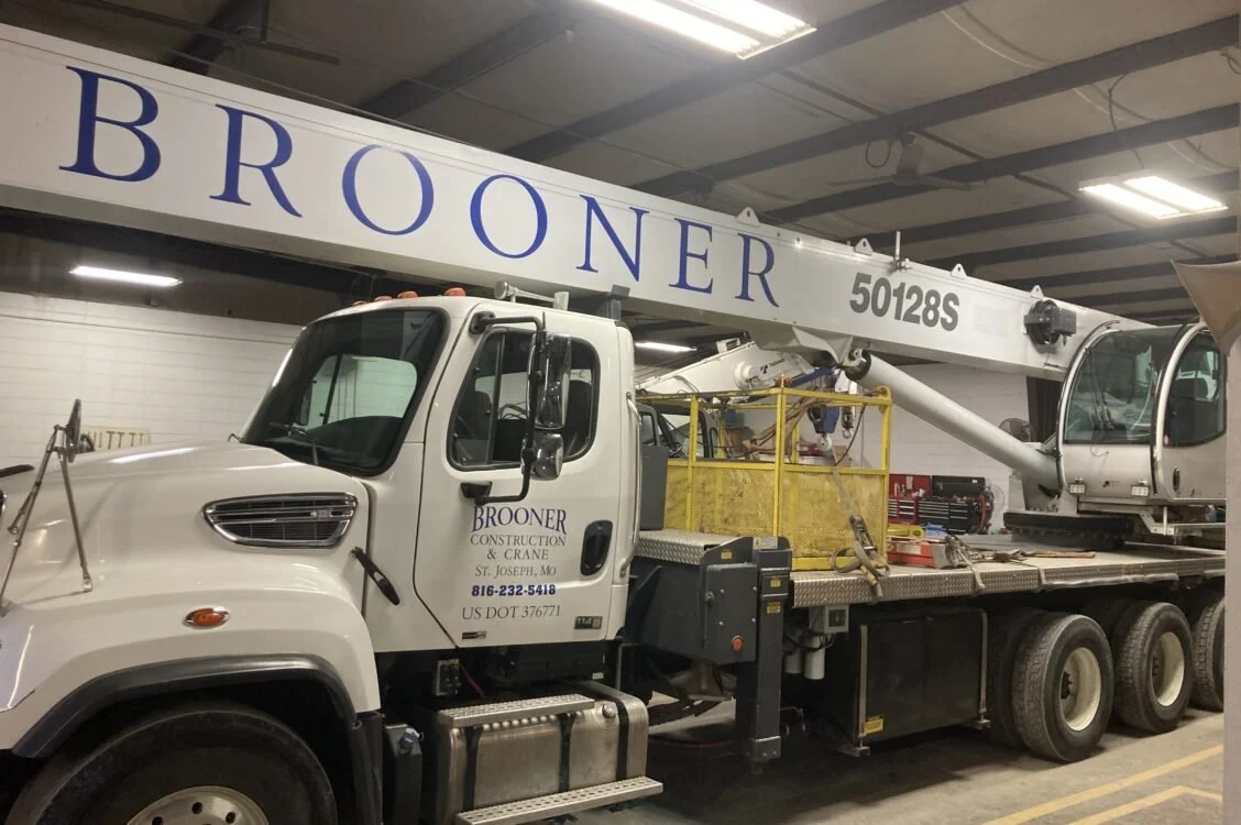 A white crane truck with branding 'BROONER CONSTRUCTION & CRANE' parked inside a warehouse. The truck has a large extendable crane labeled 'BROONER' and '50128S' on the boom.