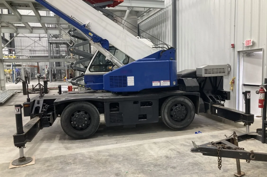 A blue and white mobile crane inside a warehouse. The crane has an extended arm with a lattice boom and is supported by stabilizer legs.