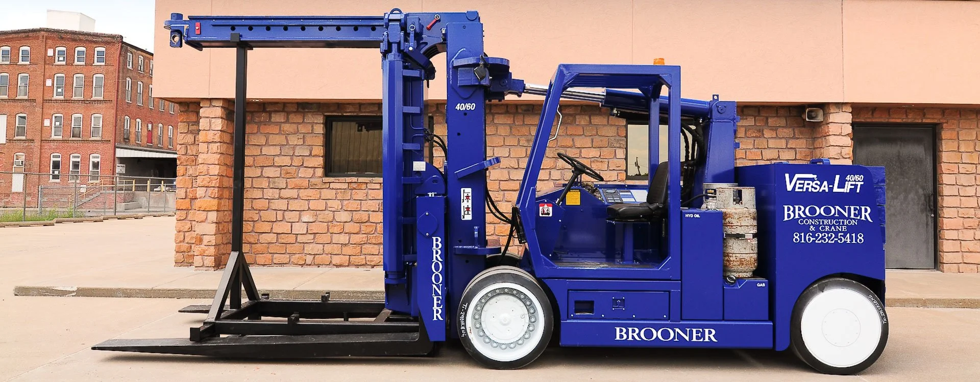 A blue forklift with branding for Brooner Construction and Crane, parked in front of a brick building with a window and a door.