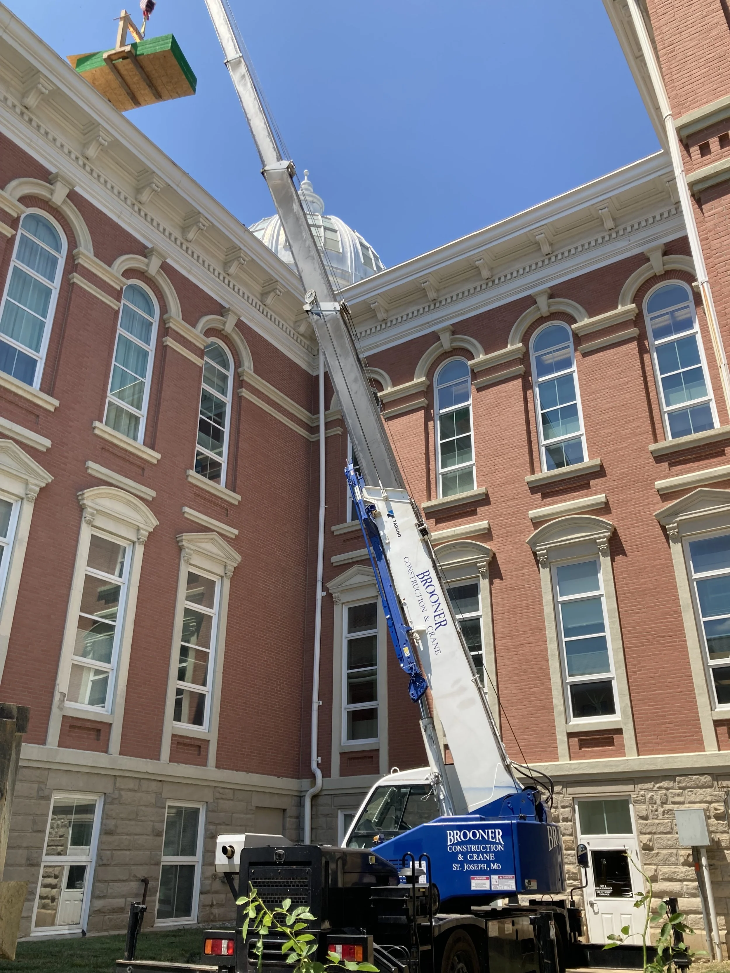 A crane labeled 'Brooner Construction & Crane' is raised near a red brick building with tall, arched windows and classical architectural details, reaching up to a clear blue sky.