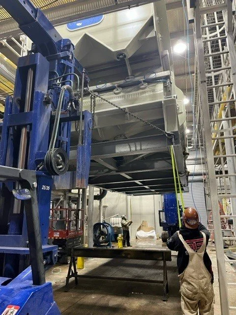Worker operating machinery inside an industrial factory. A large machine is lifted and supported with chains, with a forklift nearby.