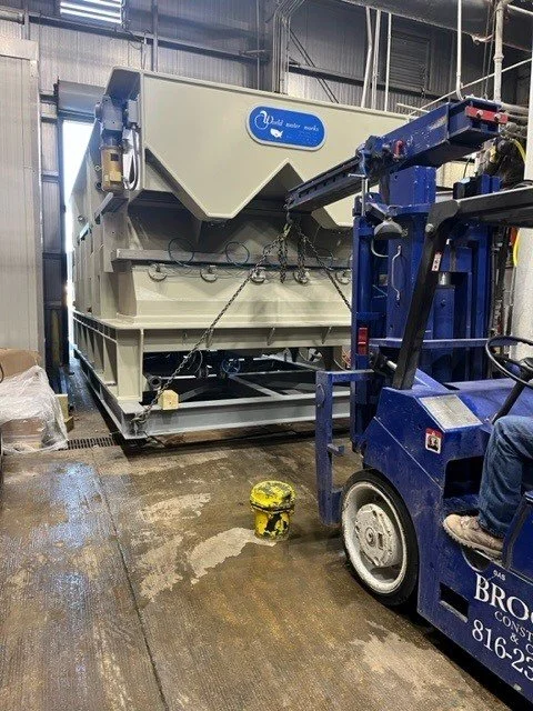 Industrial scene with large beige machine and blue forklift inside a warehouse.