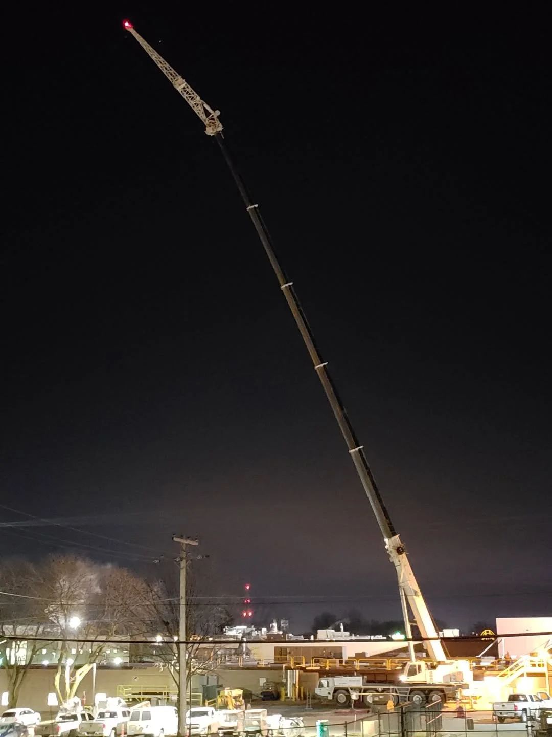 Night scene showing a large construction crane extended high into the dark sky, illuminated by construction site lights, with vehicles parked below and industrial buildings in the background.