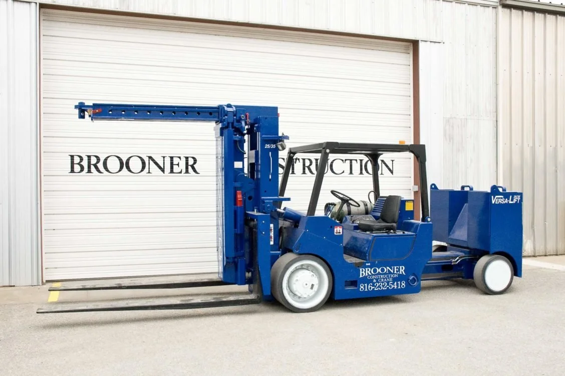Blue construction forklift in front of a white metal building with a closed garage door. The forklift has the company name and contact number on its side.
