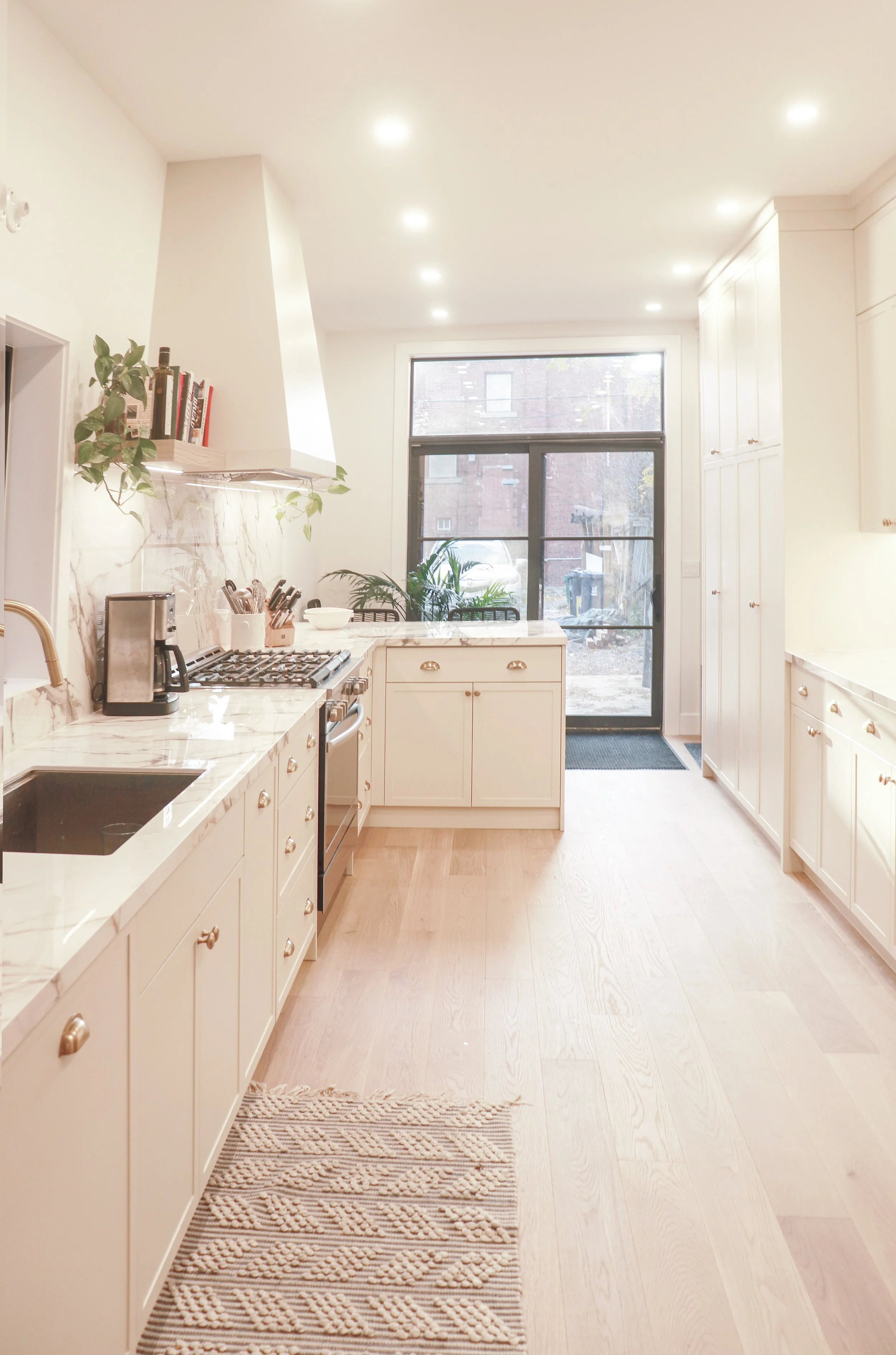 Bright, modern kitchen with white cabinets, marble countertops, a stove, coffee maker, and plants near a large window with a black frame, and a beige woven rug on light wood flooring.