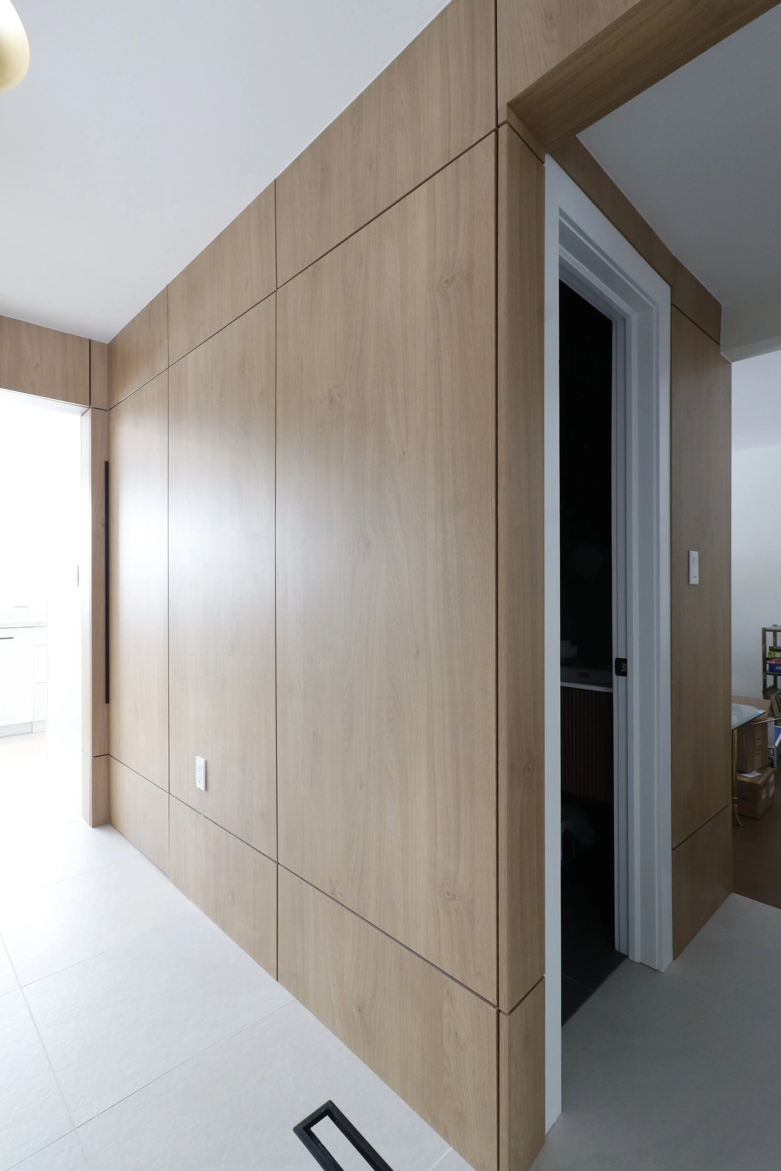 Interior view of a room with light wood paneled walls and a door leading to another room. The floor is white tile and a black floor vent is seen in the bottom left corner.