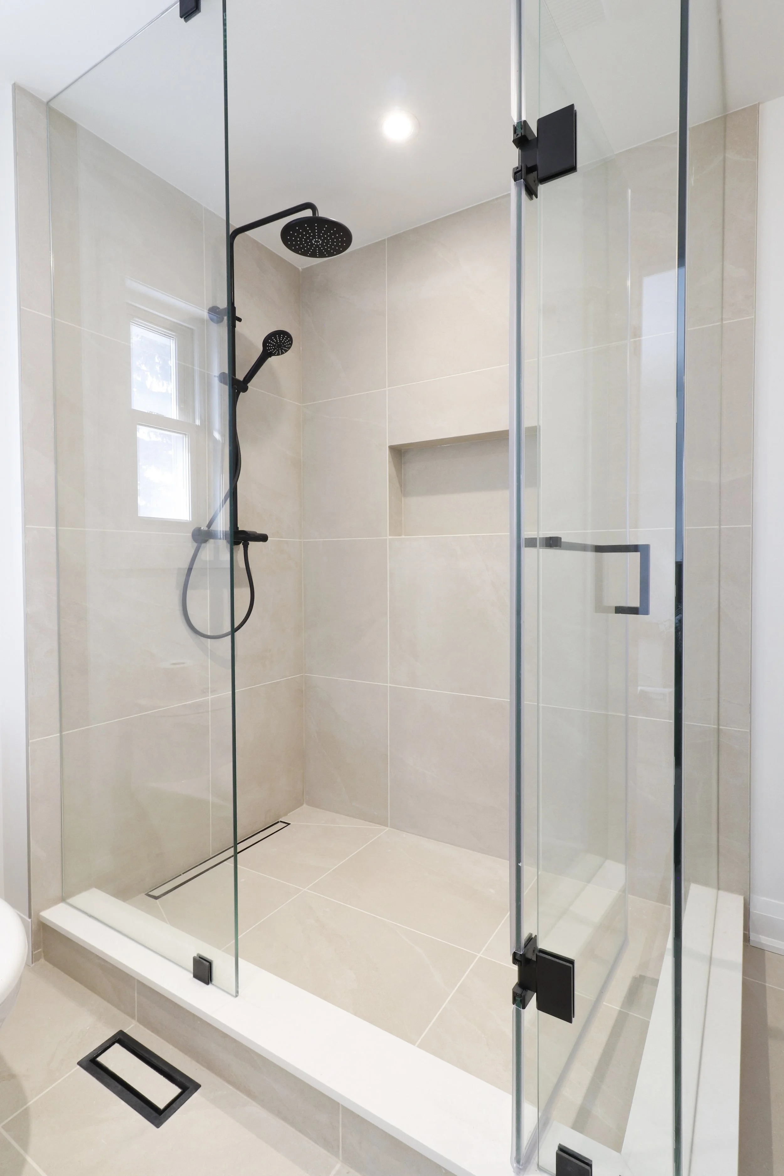 A modern walk-in shower with glass doors, black showerhead, and beige tiled walls and floor.