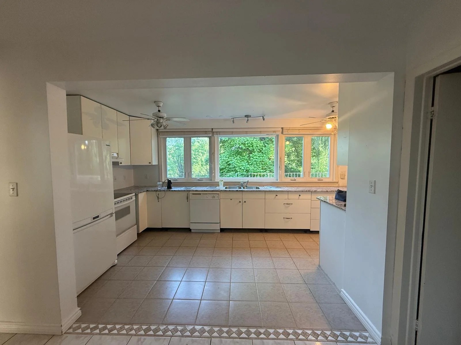 Empty kitchen with white cabinets, a white refrigerator, white oven, dishwasher, tiled floor, large window with green trees outside, ceiling fans, and track lighting.