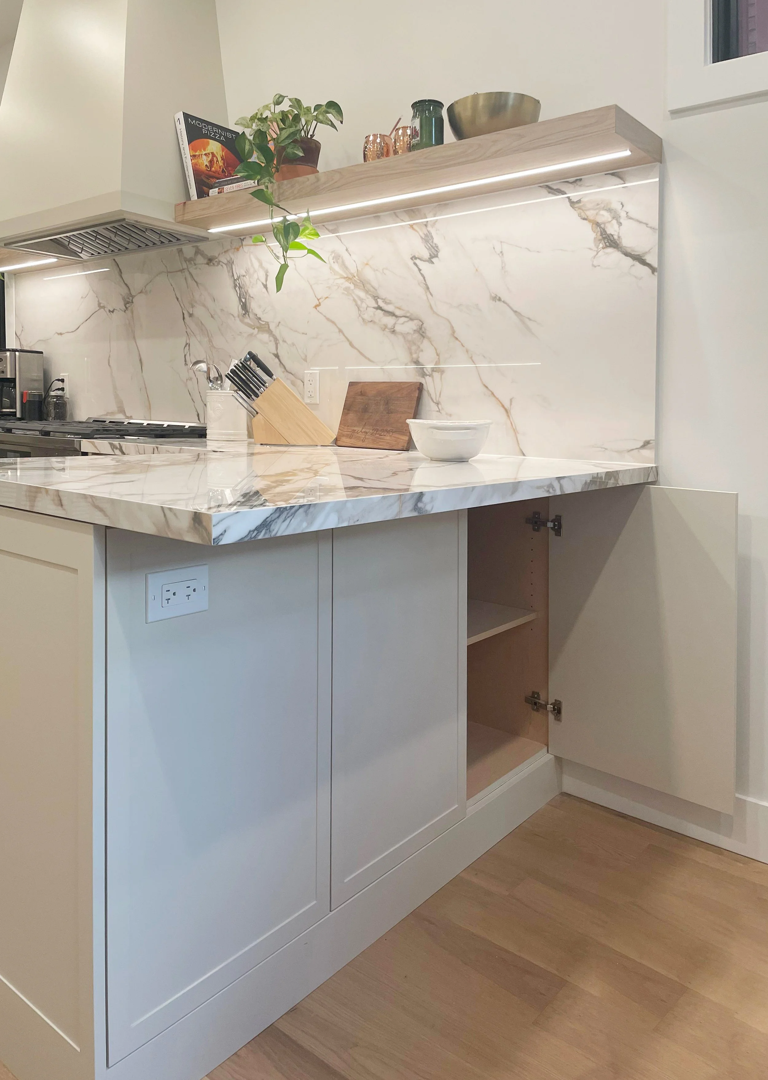 Modern kitchen with white cabinets, marble countertop and backsplash, open cabinet with empty shelves, a cutting board, bowl, and kitchen utensils.