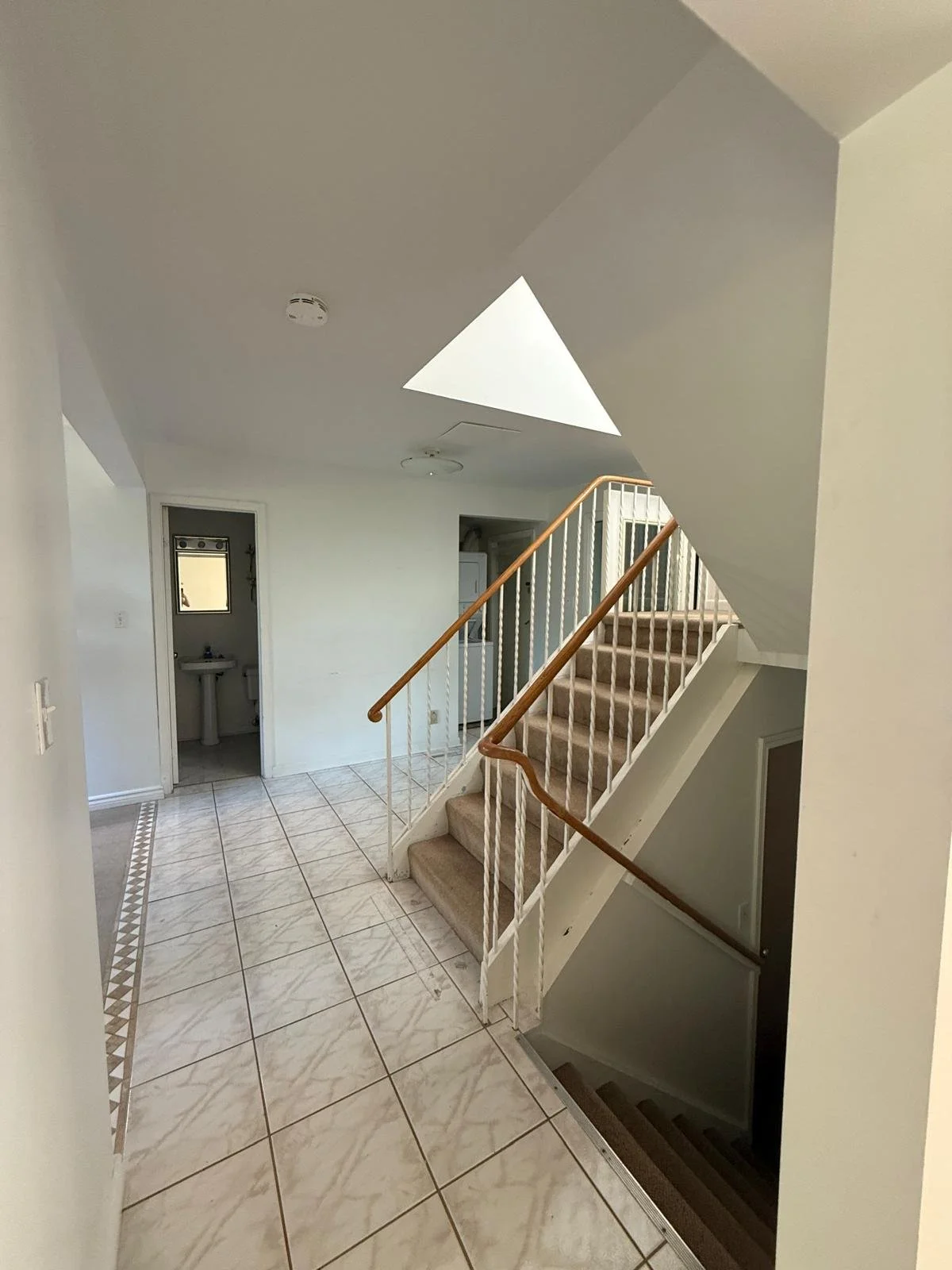 Interior view of a house entryway with tiled floor, staircase with beige carpet and wooden handrail, small bathroom with sink and toilet, skylight, and white walls.
