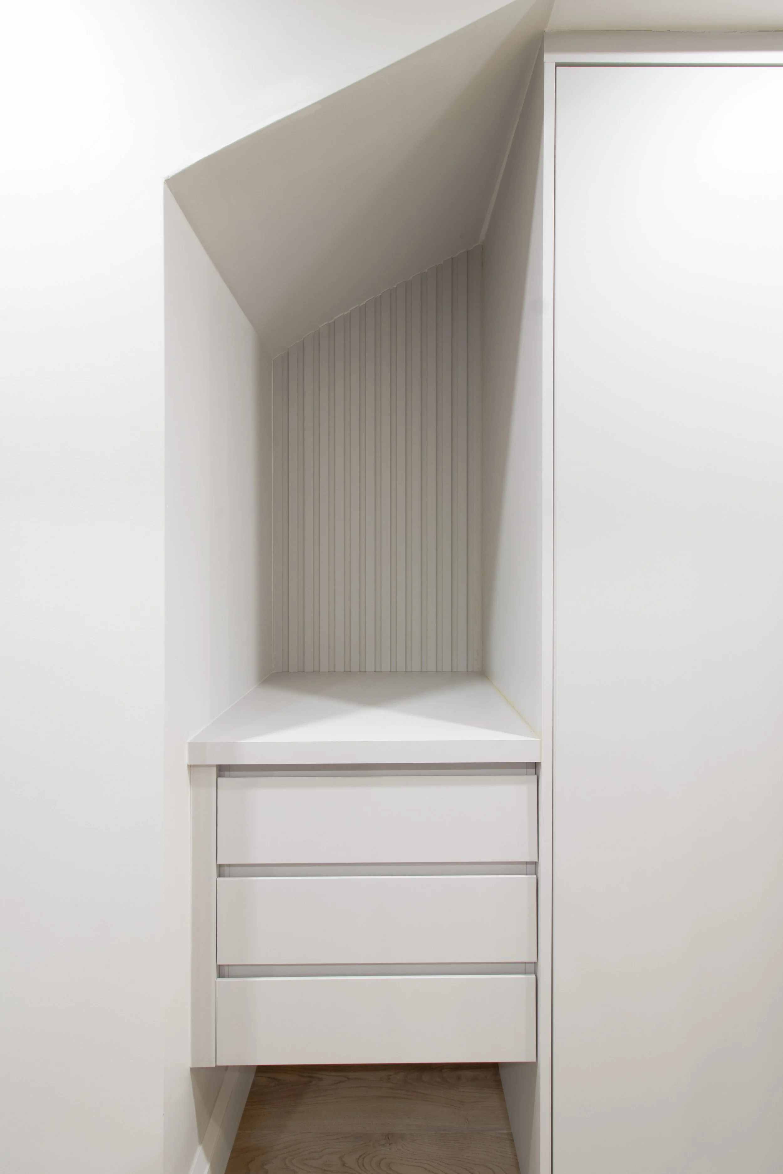White built-in cabinet with multiple shelves and drawers, set against a white wall with a sloped ceiling, and wooden flooring.