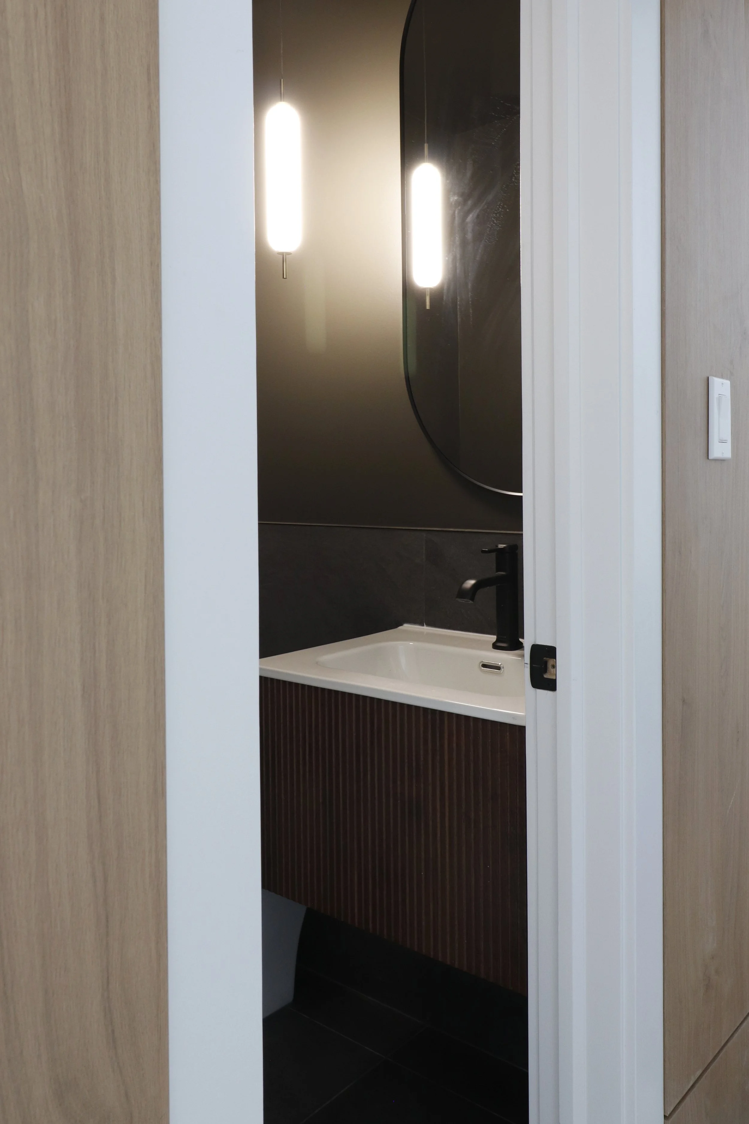 Bathroom with black sink and faucet, wall with black and brown textured tiles, mirror, and hanging lights, partially visible through a doorway