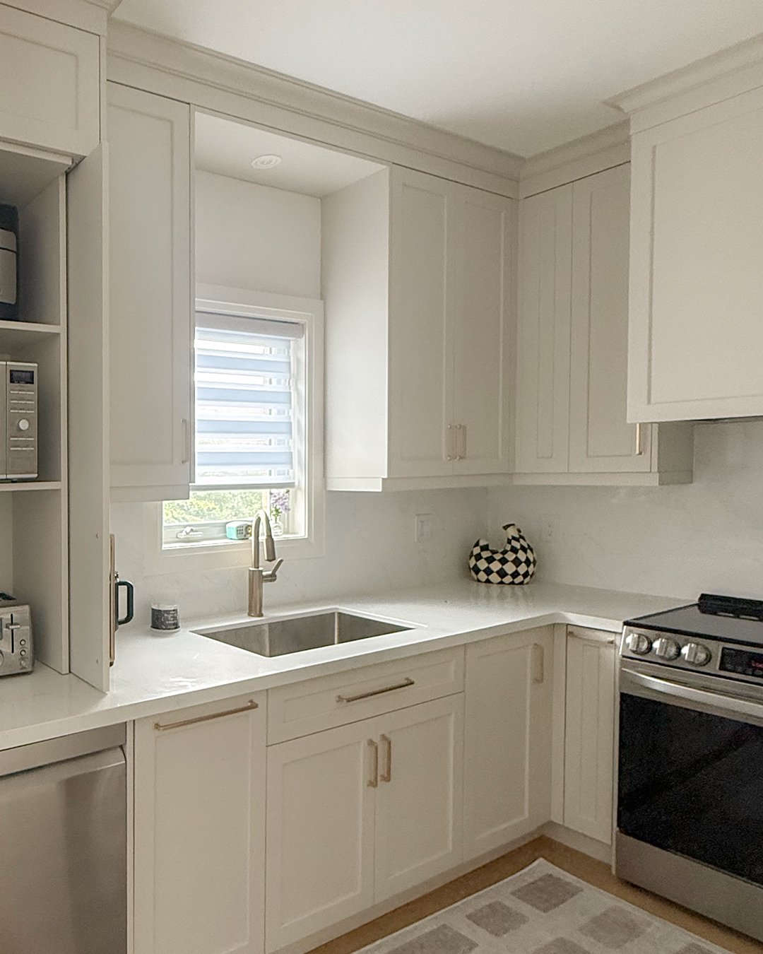 White kitchen with upper and lower cabinets, a small window above the sink, a black-and-white checkered decorative item on the countertop, a stove, and various small appliances on the shelves.