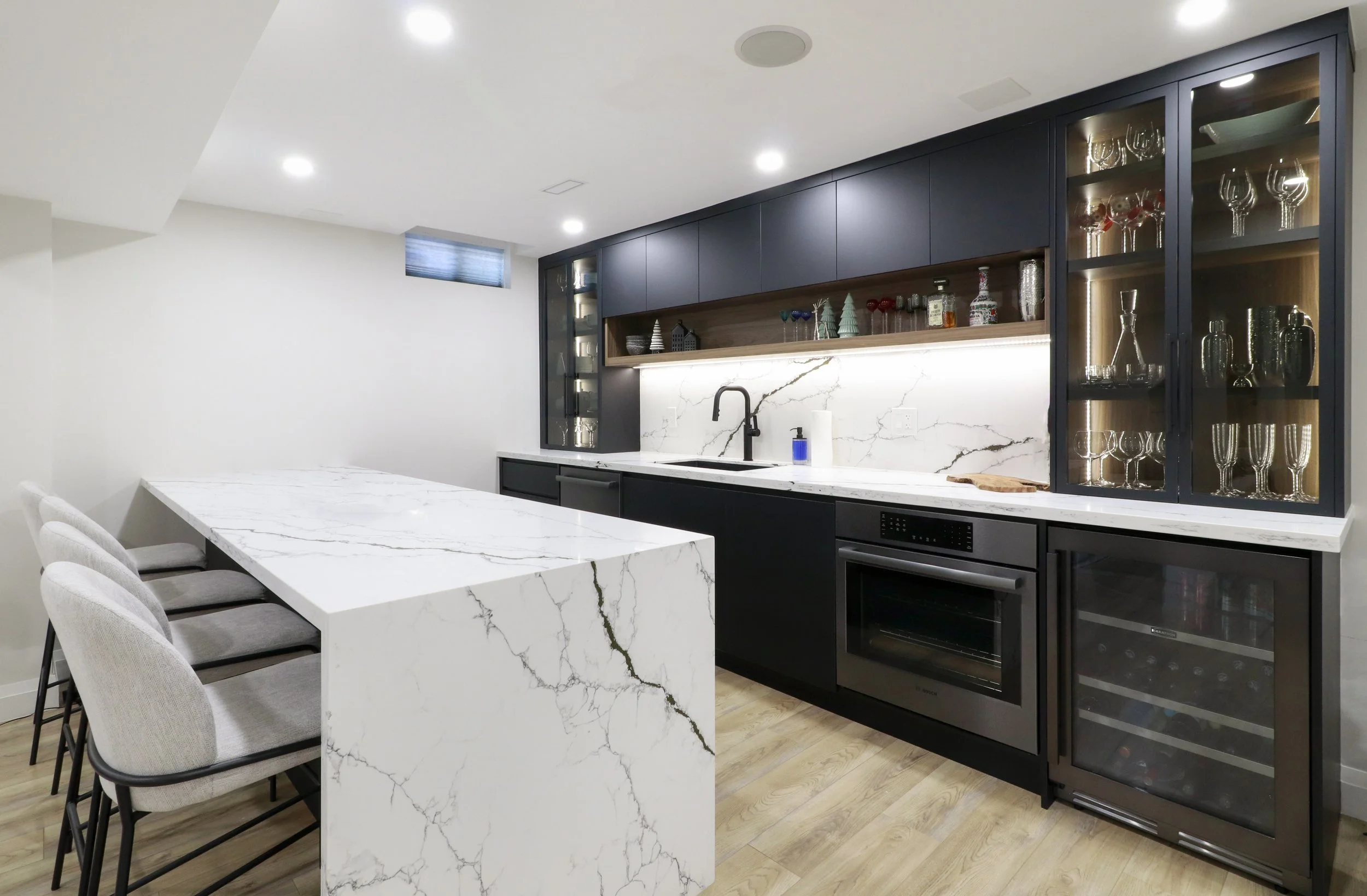 Modern kitchen with a white marble island, black cabinetry, and glass-fronted cabinets displaying glassware. Recessed ceiling lights and a small window are visible.