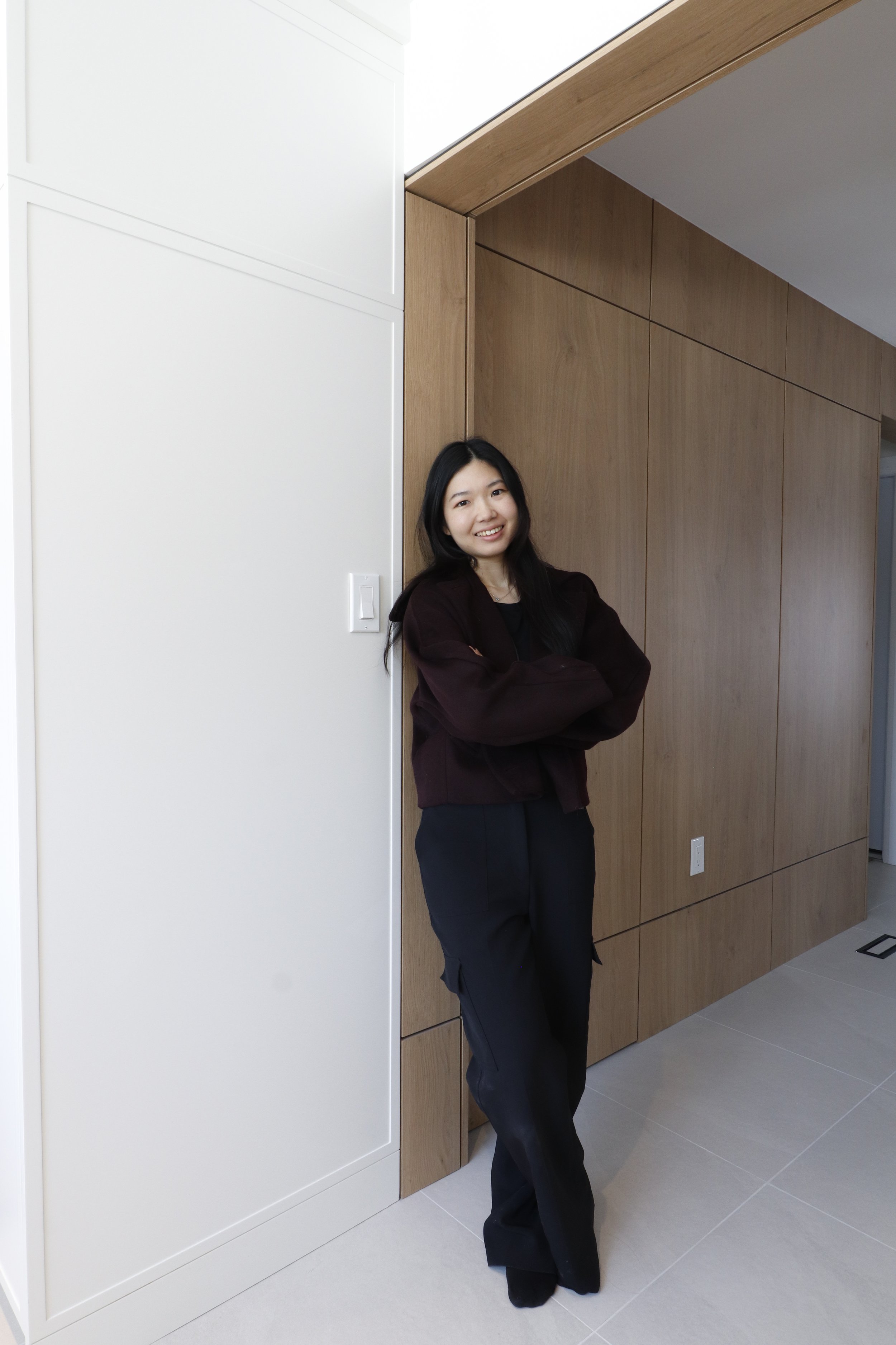 A woman with long black hair smiling and crossing her arms, standing inside a modern room with wooden panel walls and a white door.