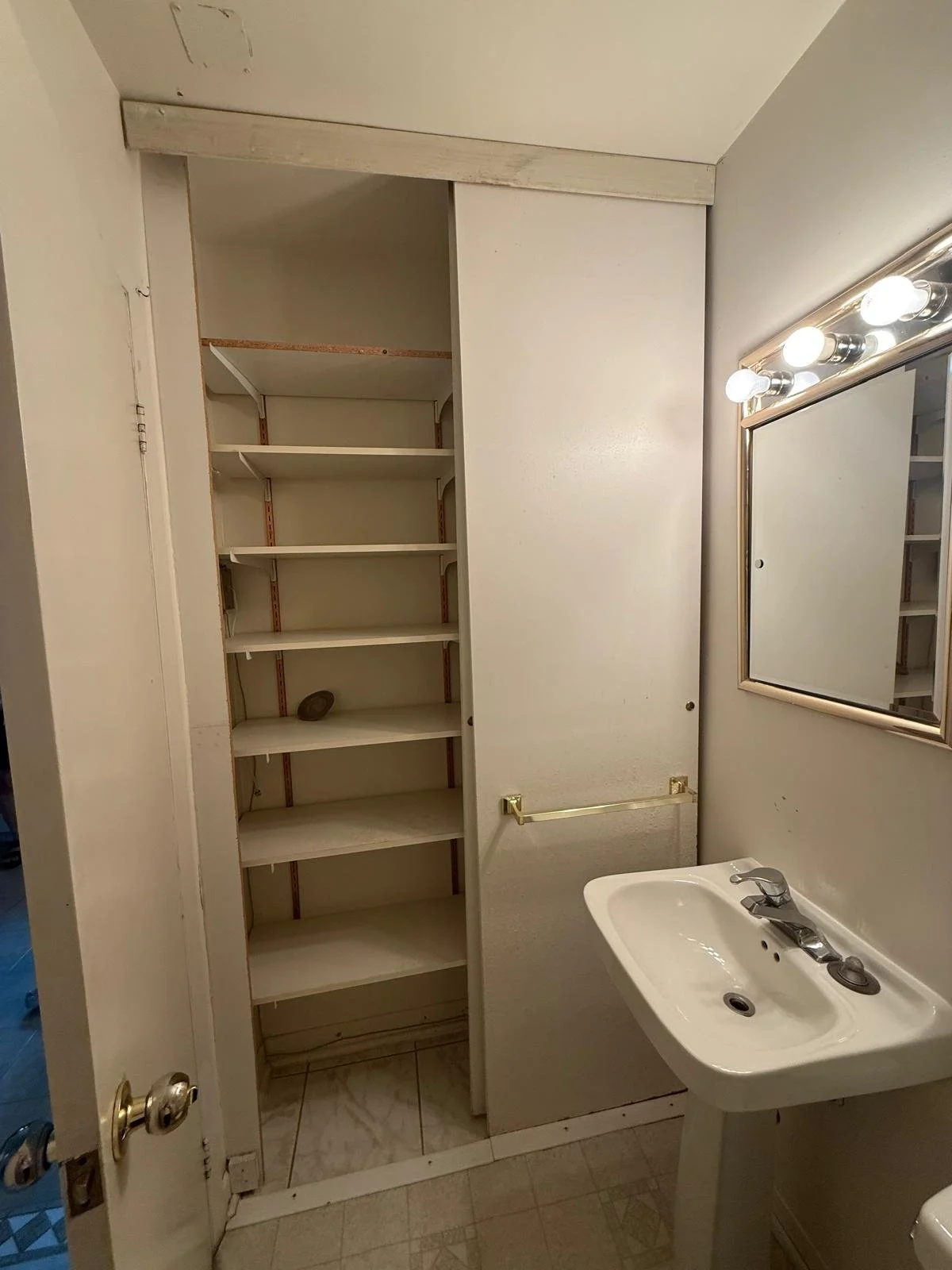 A small bathroom with a white sink, a mirror with three light bulbs above, a partially open cabinet with shelves, and a towel bar. The bathroom has beige tile on the floor and a wall with a sliding door. The shelves inside the cabinet are mostly empt