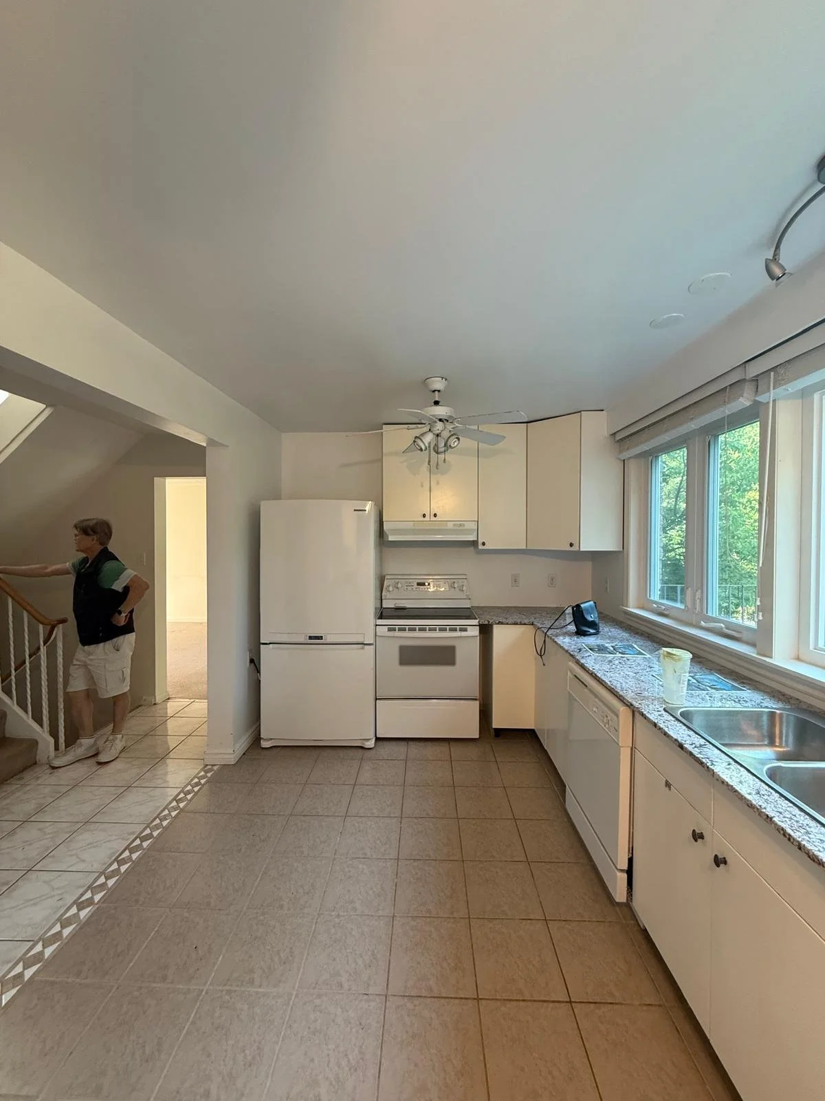 Kitchen with white appliances, granite countertops, large window, and a person standing near the staircase.