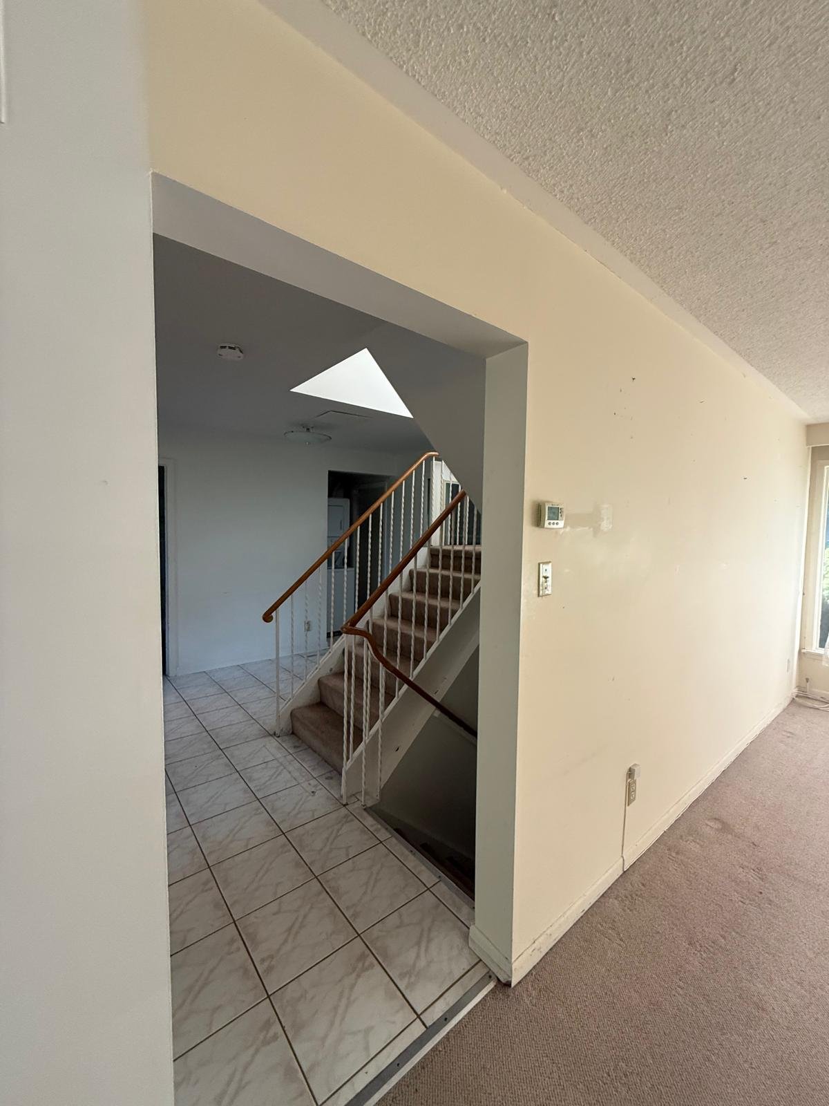 Interior view of a house showing entrance foyer with tiled floor, staircase with carpeted steps and white railing, and a wall with a thermostat and a switch. Beige carpeted area on the right.