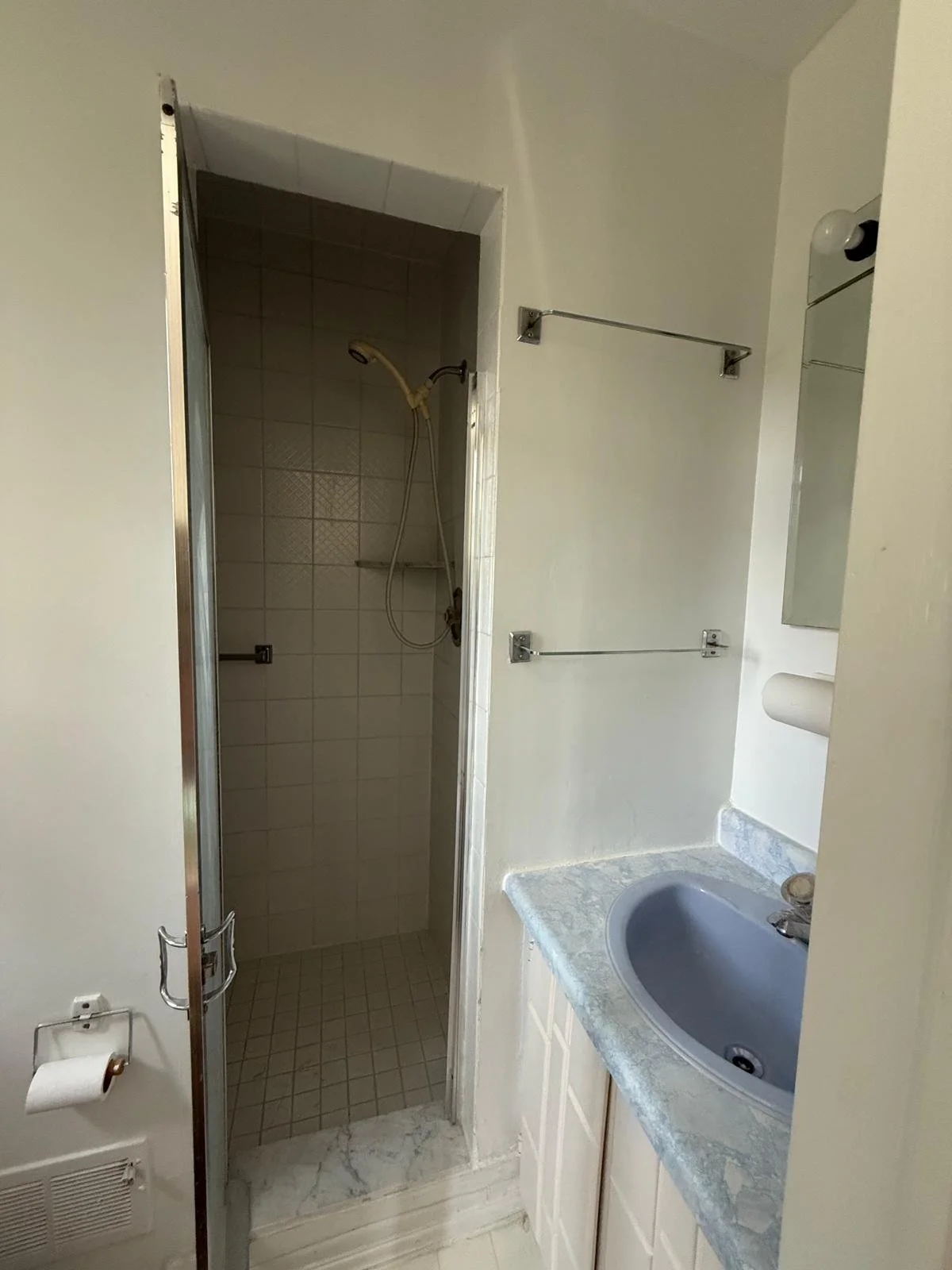 Small bathroom with a shower area and a sink with a marble countertop. The shower has white tiles and a handheld showerhead. There are two metal towel bars on the wall, a mirror above the sink, a toilet paper holder, and a partially visible mirror ca