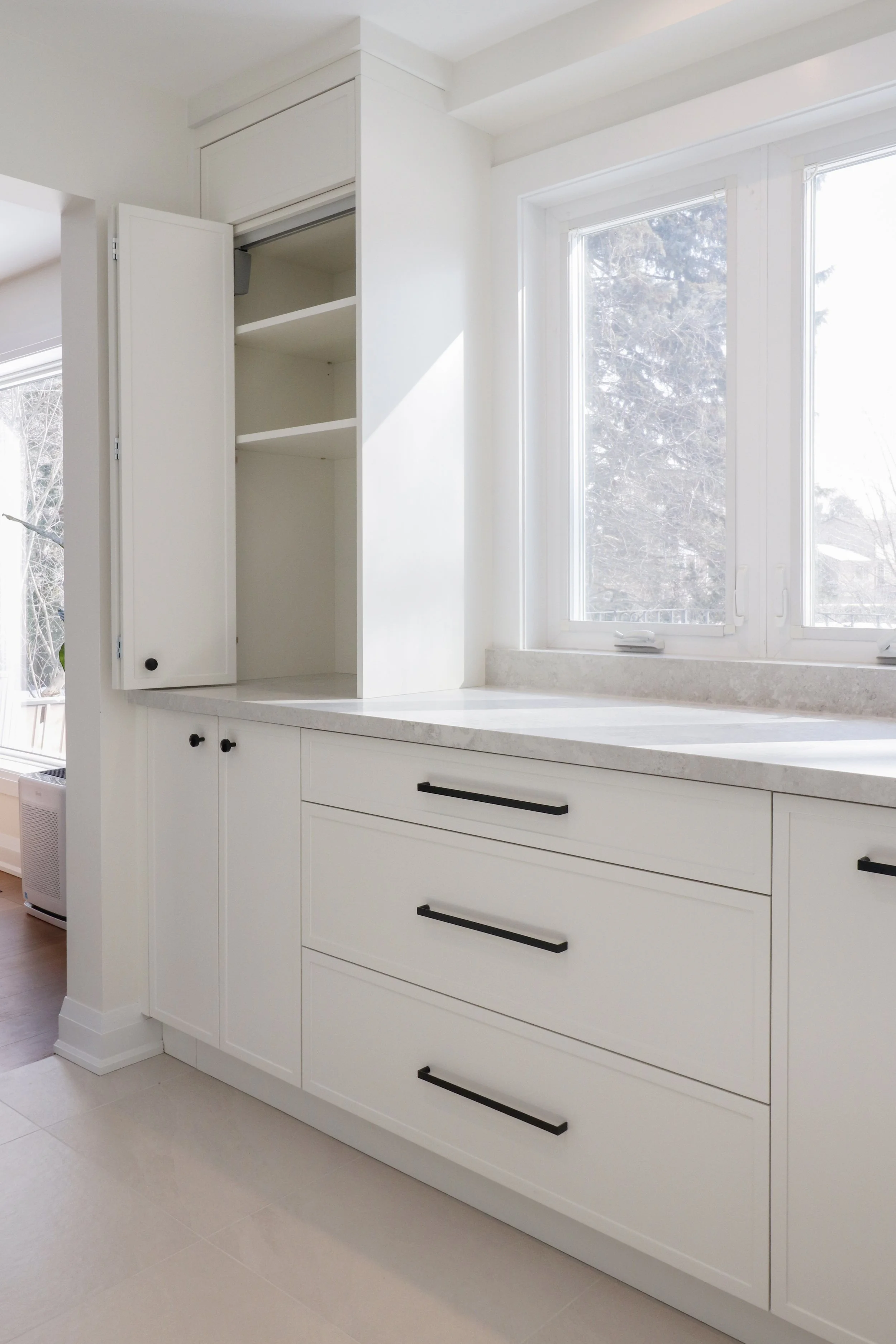White kitchen cabinet with black handles beneath a large window showing trees outside, with white countertops and a beige floor.