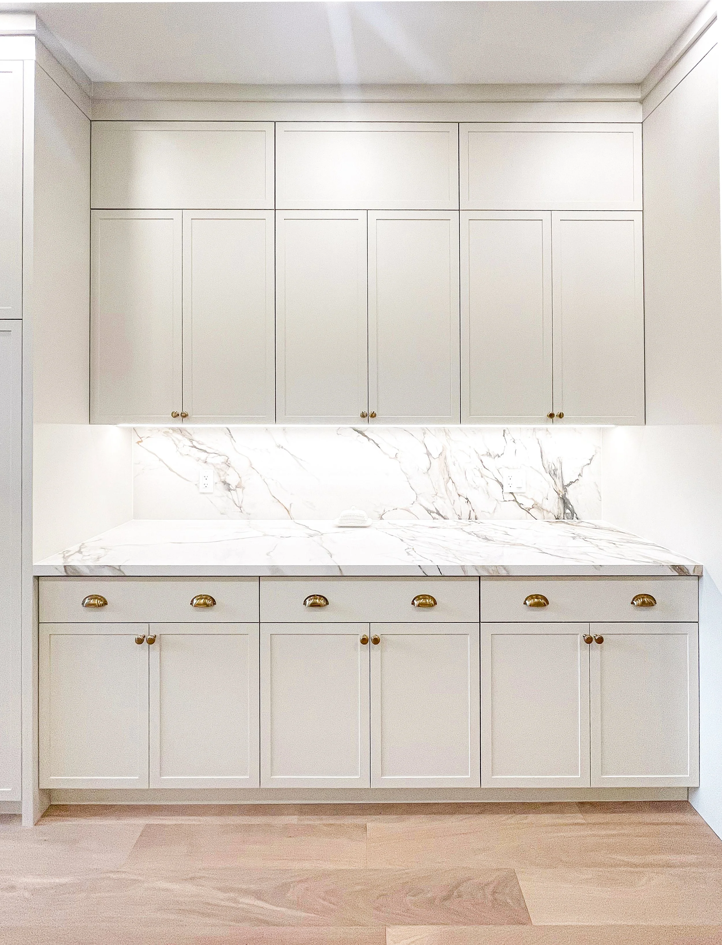 White kitchen cabinets with gold knobs, marble countertop and backsplash, and light wooden flooring.