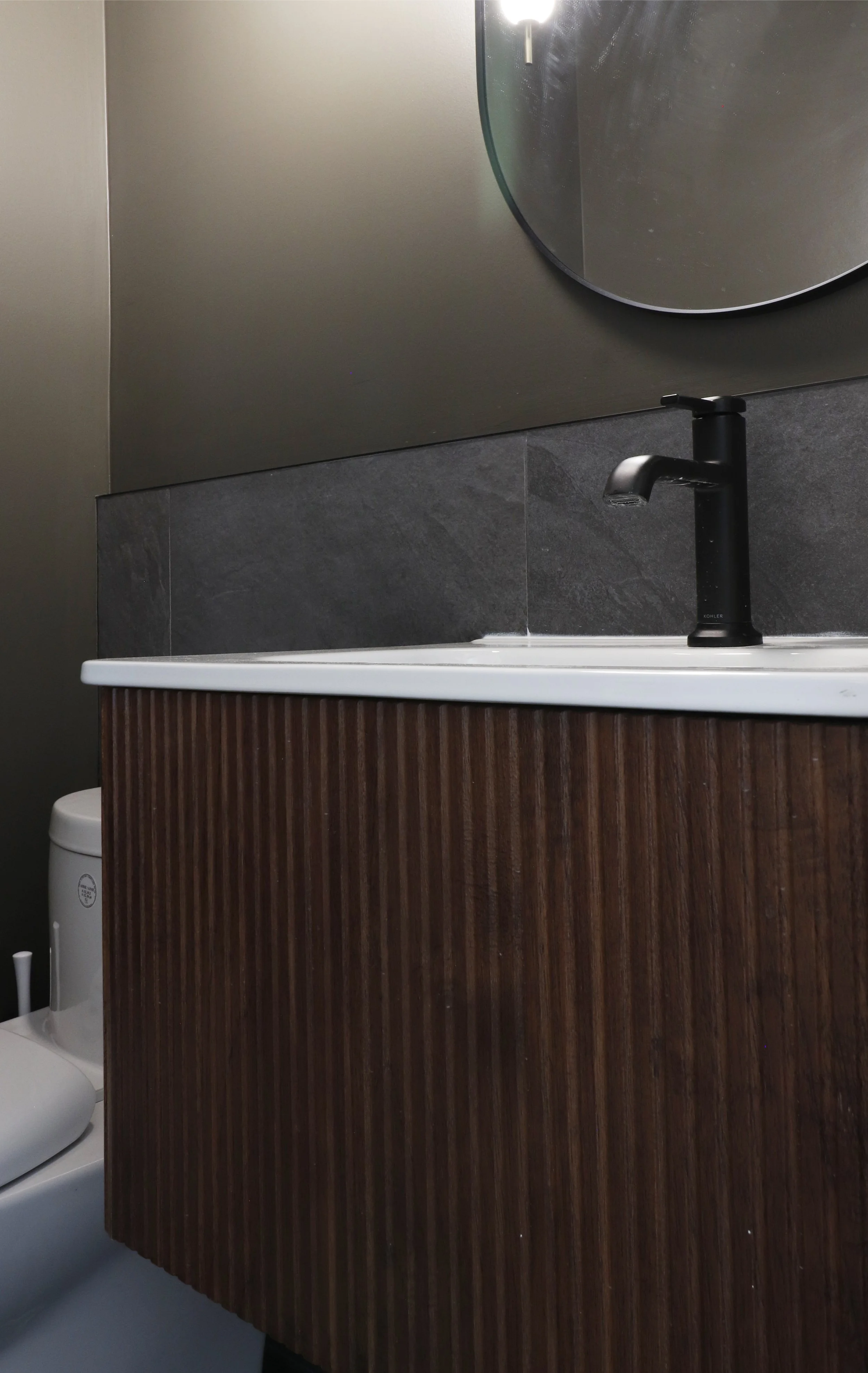 Modern bathroom sink with black faucet on a wooden vanity, mirror on dark gray wall, and part of a white toilet visible on the left.