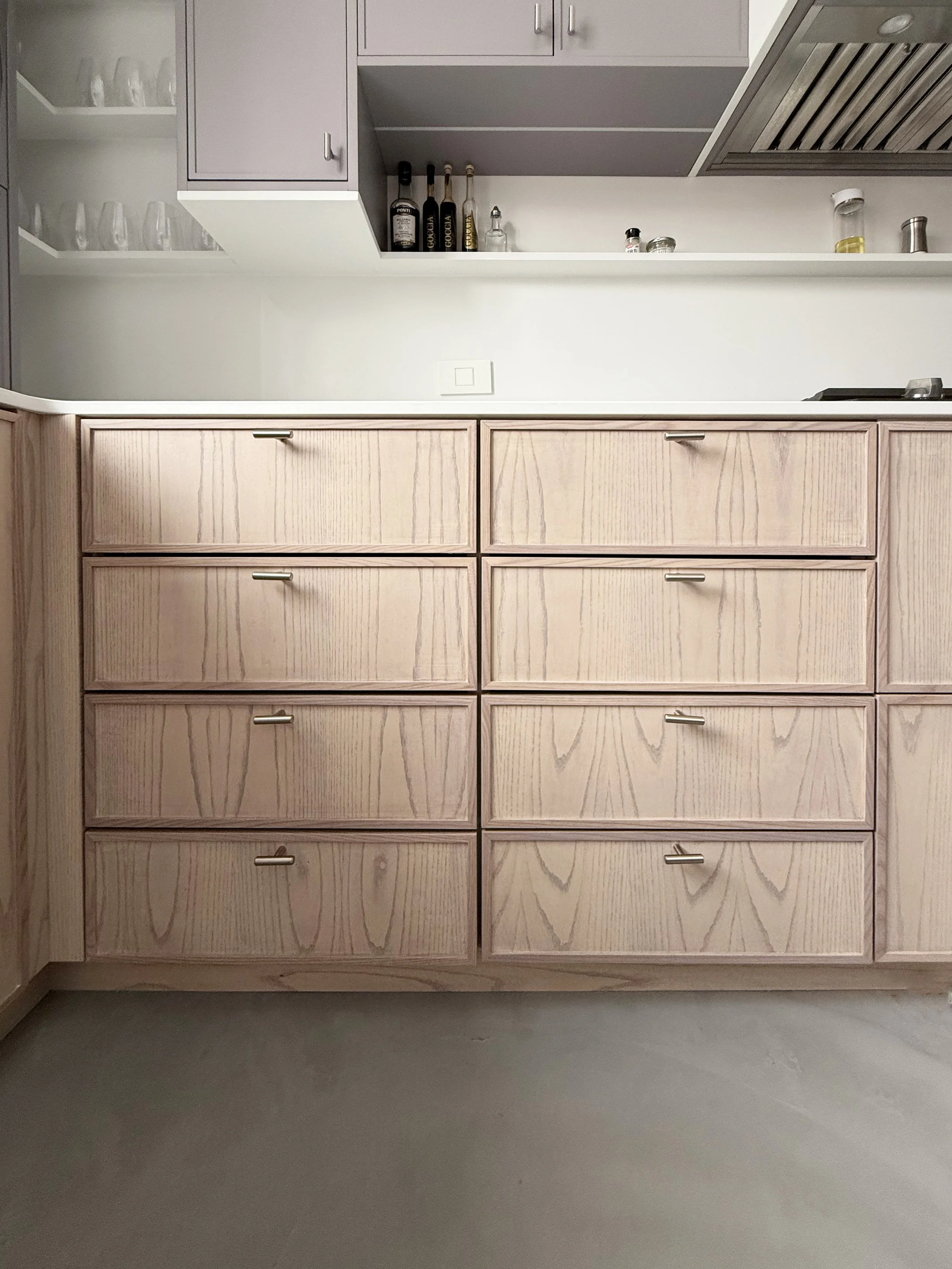 View of a kitchen with a light-colored wooden cabinet with ten drawers and a white countertop. Upper cabinets in soft gray and a white open shelf with glasses and various bottles. The backsplash is white, and there is a stainless steel range hood. Th