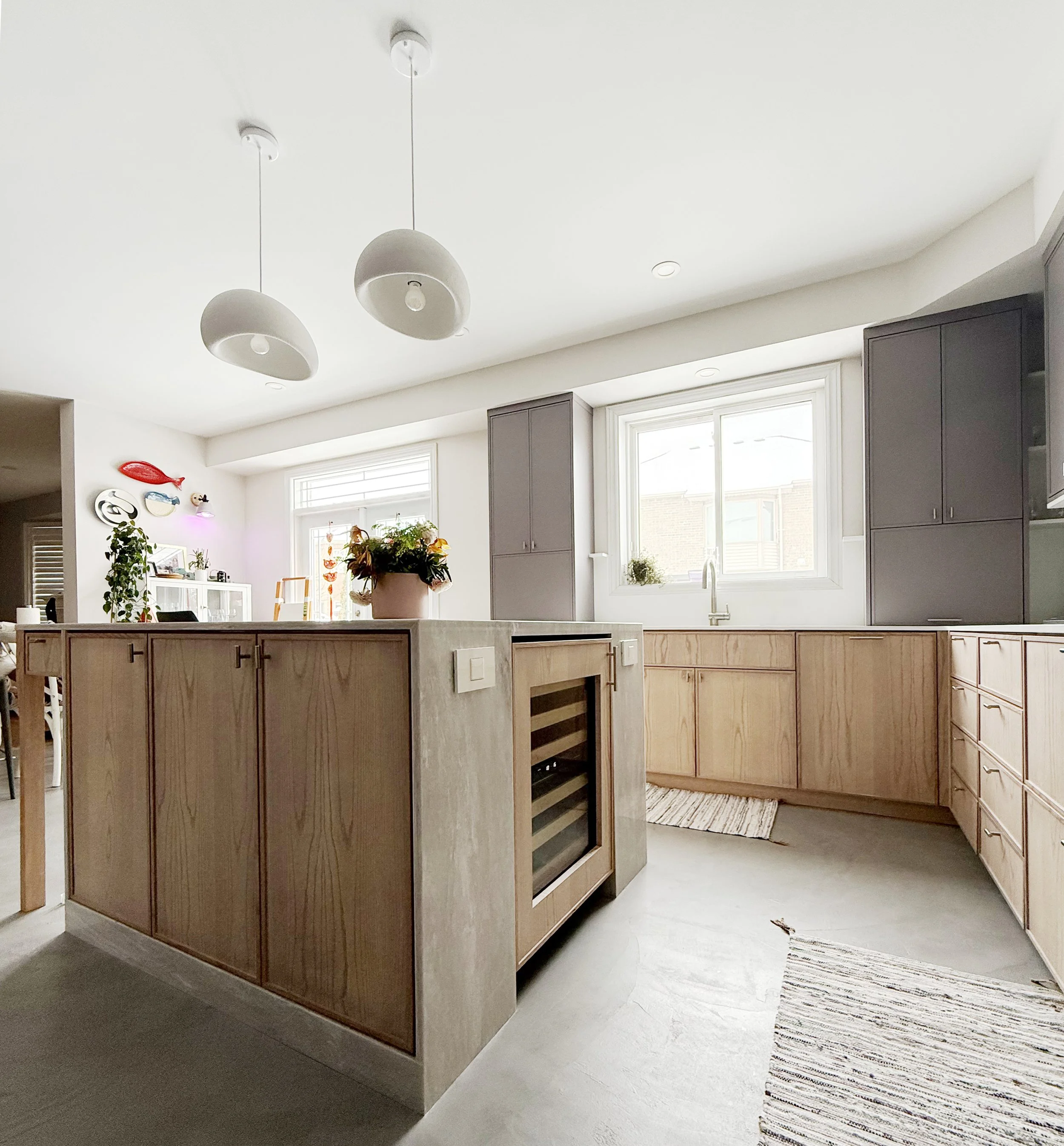 View of a room with a kitchen island, gray kitchen cabinets, a window, and a striped rug on a concrete floor. Decor includes plants and wall art, with ceiling lights.