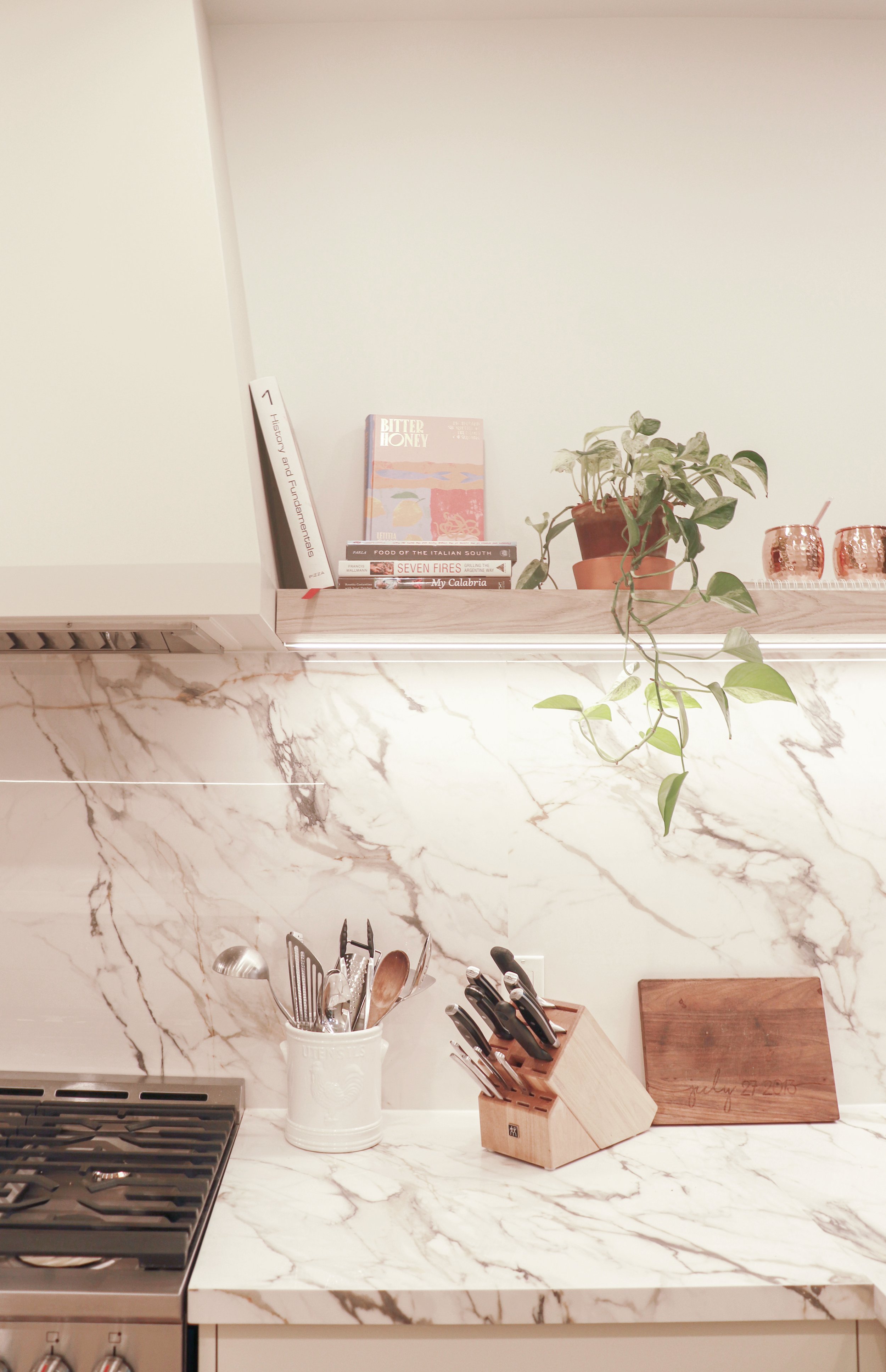 Kitchen counter with a marble countertop, a gas stove, a white utensil holder with utensils, a wooden knife block with knives, a wooden cutting board, a potted plant, and shelves with books and decorative items.