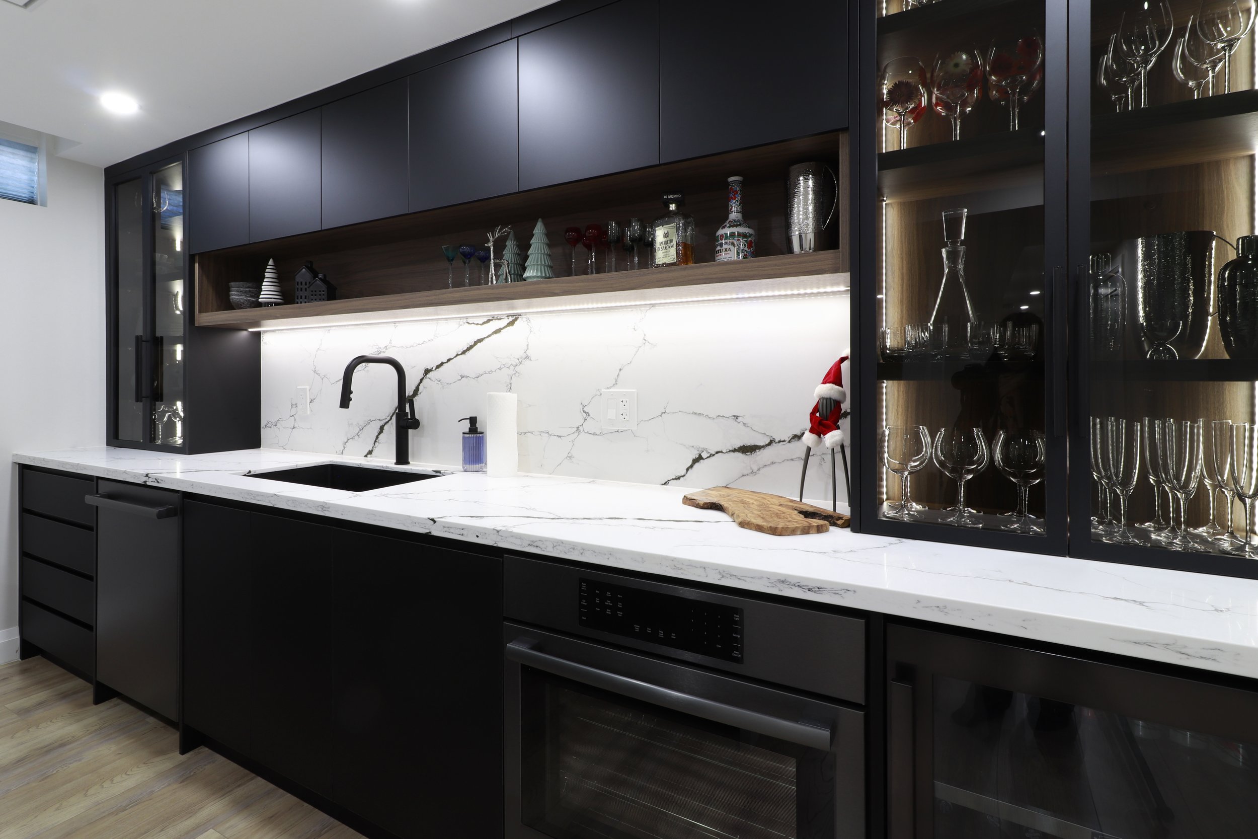 Modern kitchen with black cabinets, white marble countertop, and a marble backsplash. Glassware displayed in a black cabinet with a wooden interior. Decor includes small Christmas figurines and bottles of alcohol.