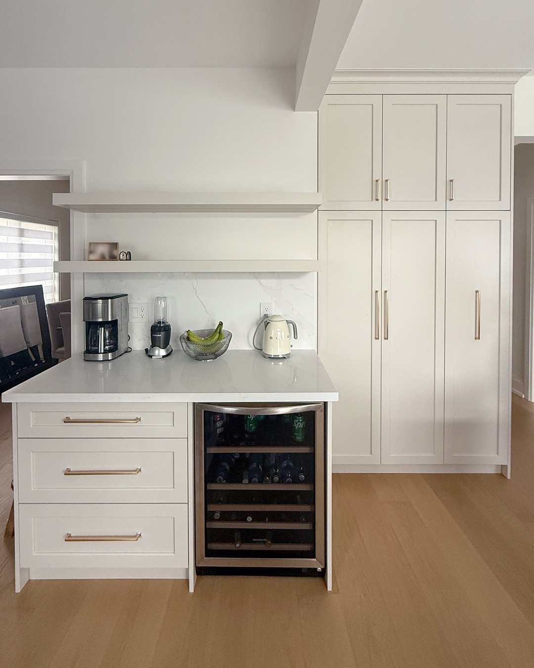 White kitchen counter with a wine cooler, a coffee maker, a blender, a banana bowl, and an electric kettle, with white cabinets and wooden flooring.