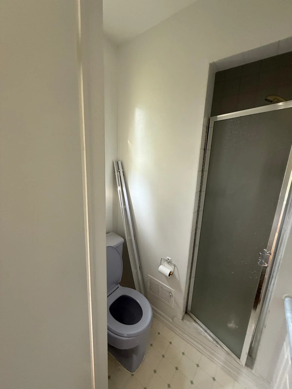 Small bathroom with a toilet, a partially opened sliding shower door, and a wall-mounted toilet paper dispenser. There are two metal rails leaning against the wall near the toilet.