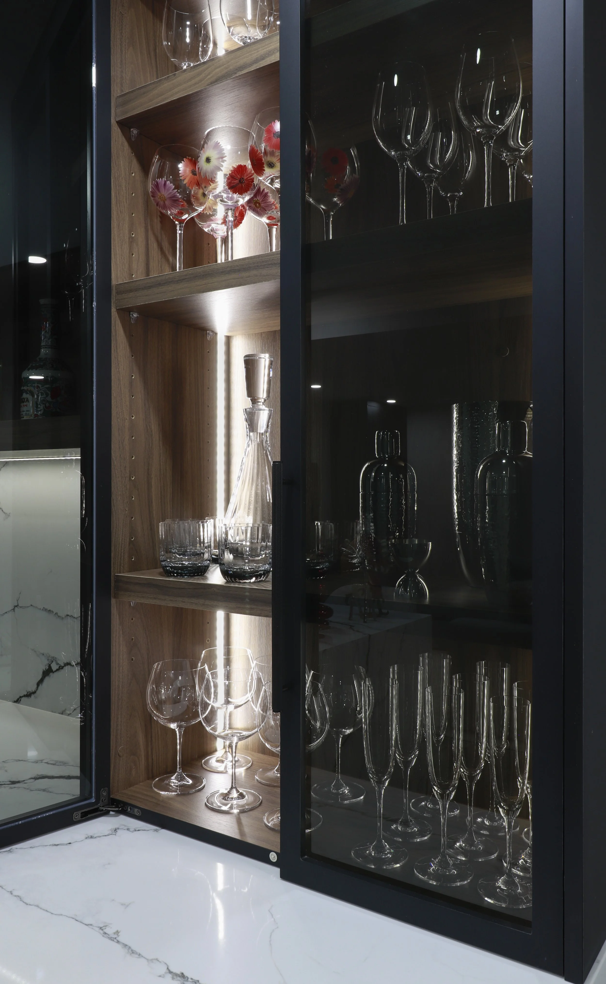 Glass cabinet with wooden shelves holding various glassware including wine glasses, champagne flutes, and decanters, some with floral designs and others plain, on a marble countertop.