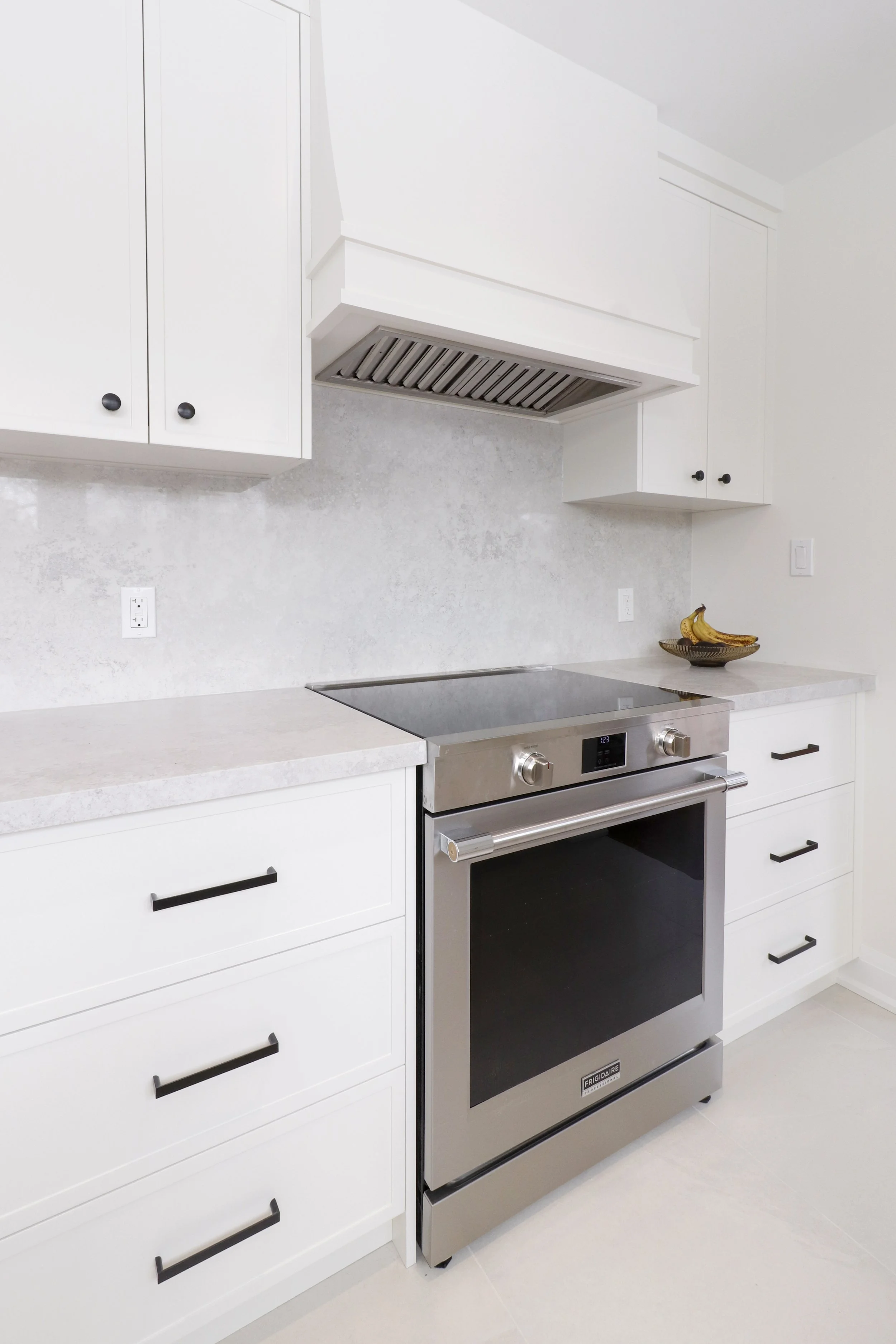White modern kitchen with white cabinets, black handles, a stainless steel oven, a white countertop, a banana in a black bowl, and a wall-mounted range hood.
